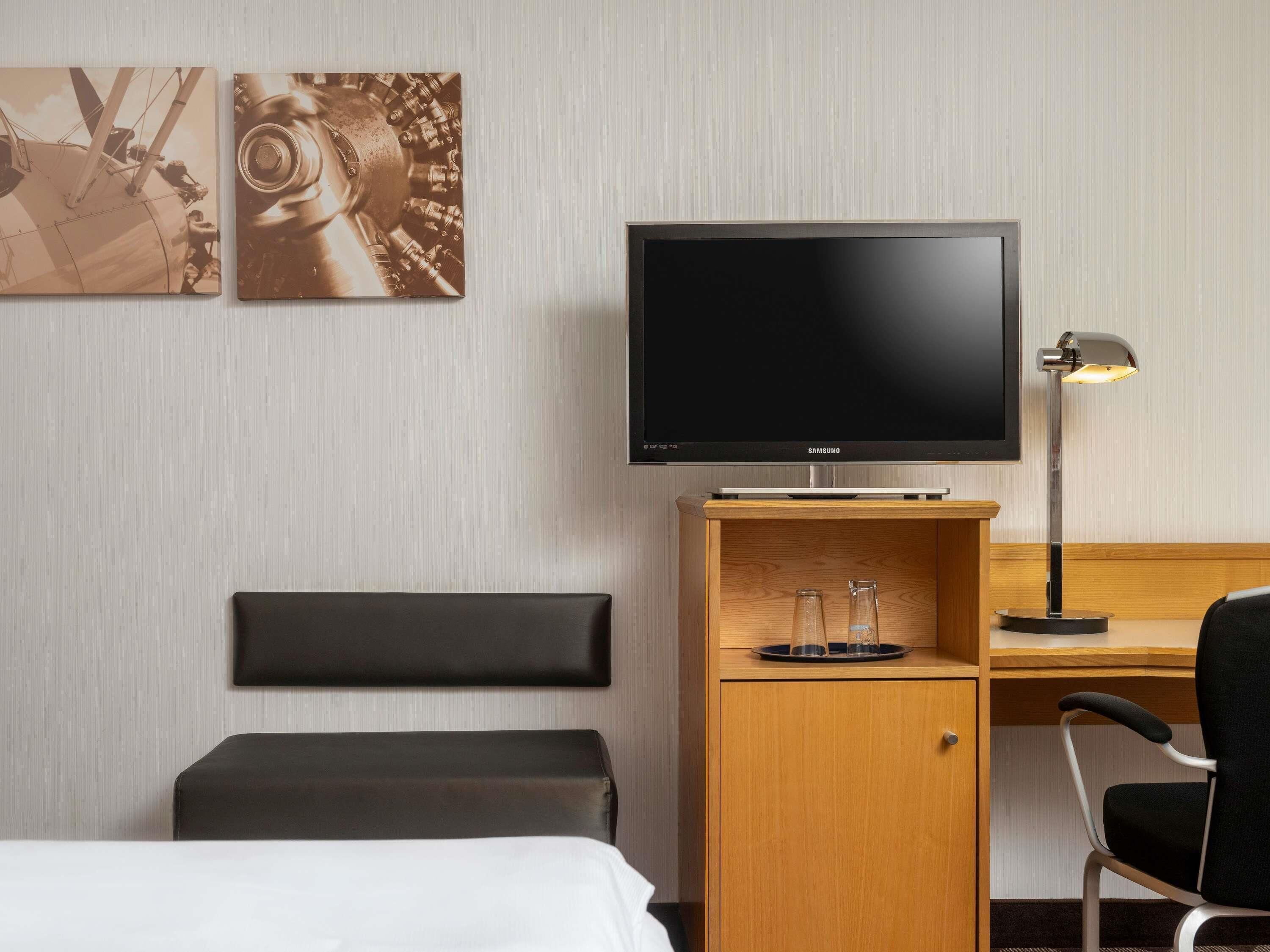 Ein Hotelzimmer mit Bett, Schreibtisch mit Fernseher, Lampe und zwei an der Wand befestigten Kunstwerken.