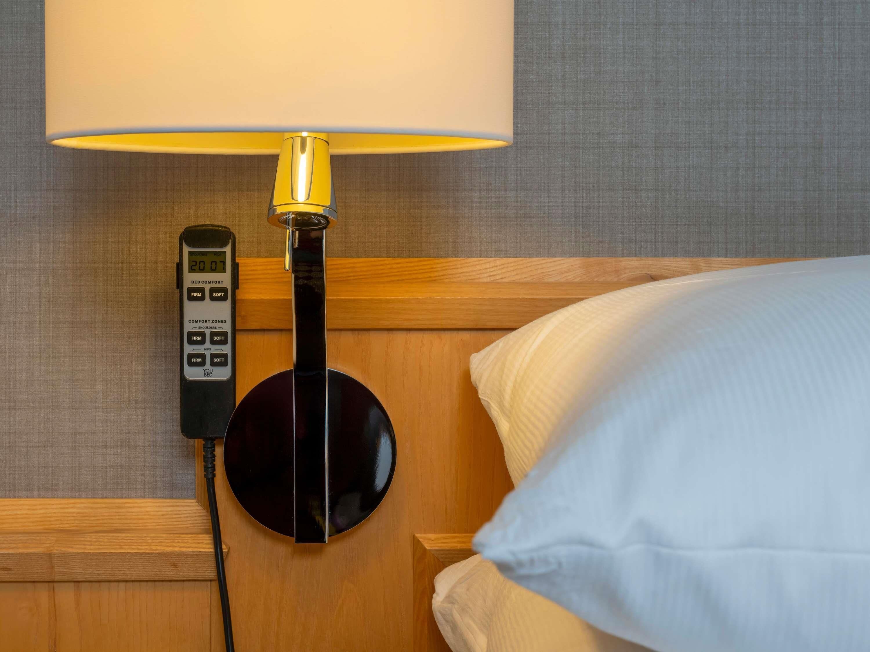 A bed with a white pillow, a bedside lamp, and a control panel on the wall.