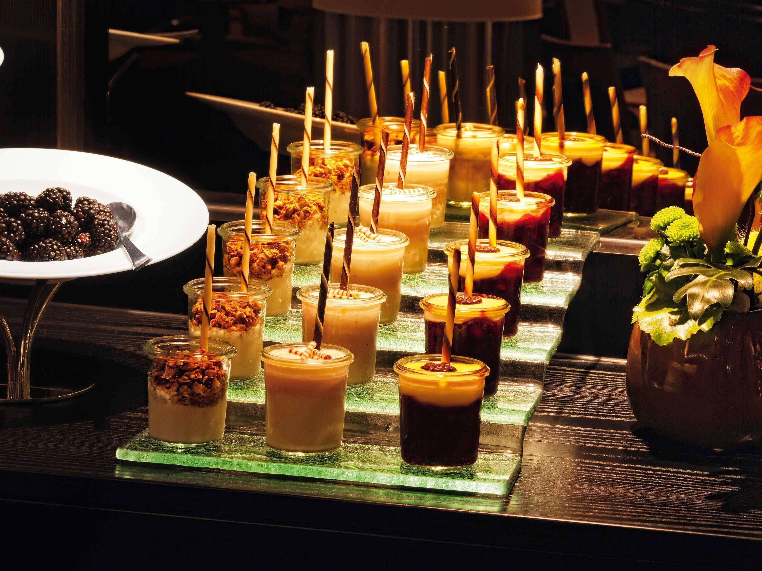 A variety of drinks in glass jars with straws, a plate of blackberries, and a vase with flowers.