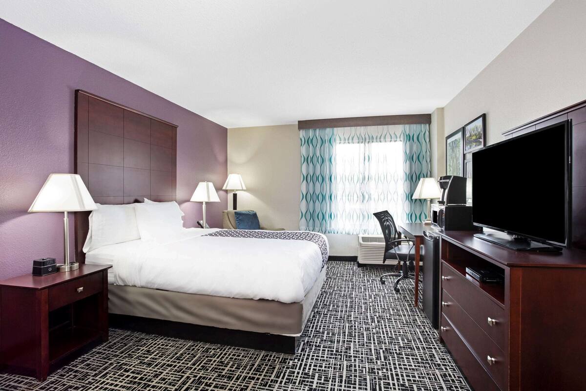 A hotel room with a large bed, two bedside tables with lamps, a flat-screen TV, a desk with a chair, and a patterned carpet.