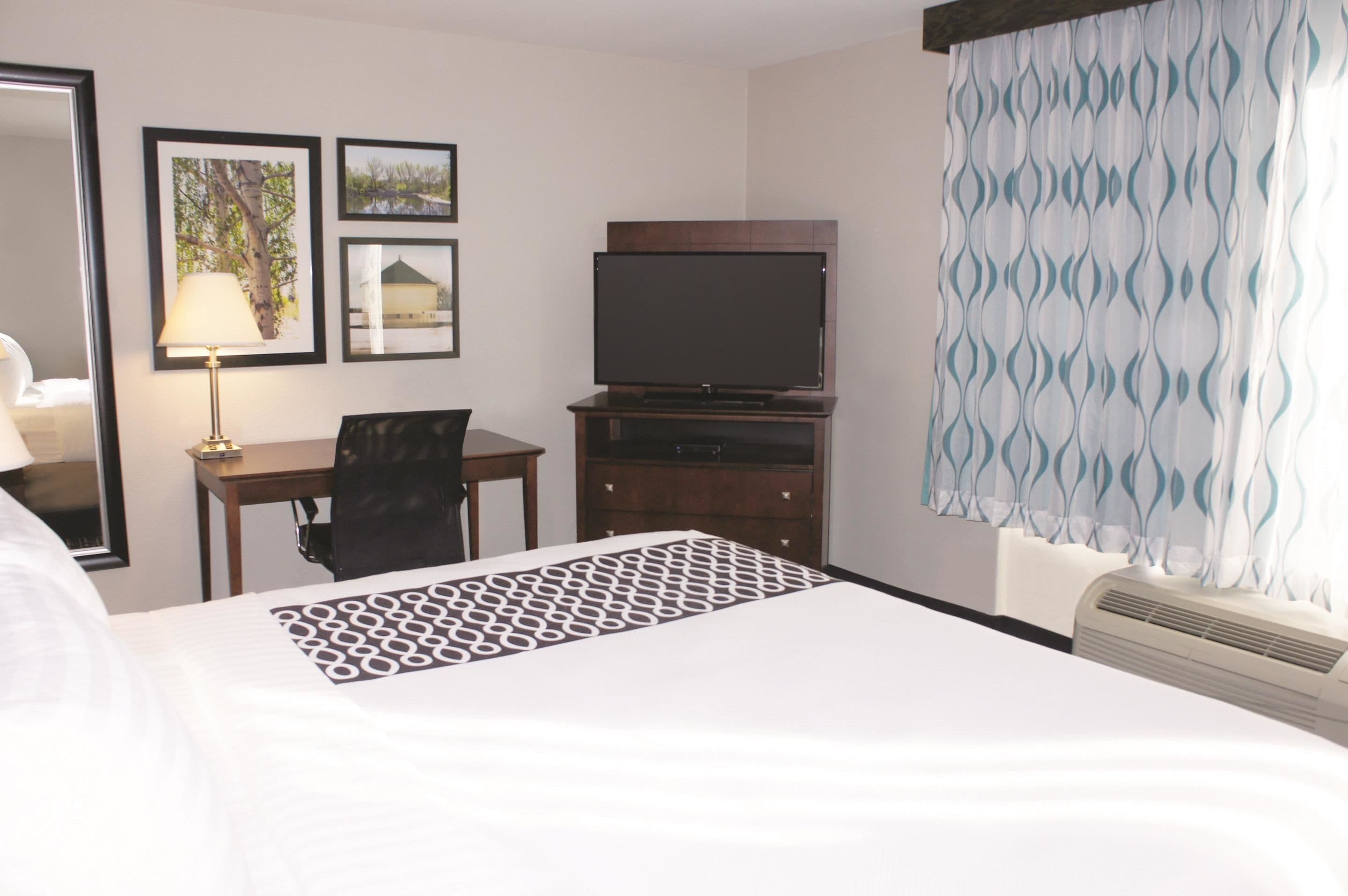 A hotel room with a bed, a desk with a chair, a television on a stand, and a window with patterned curtains.