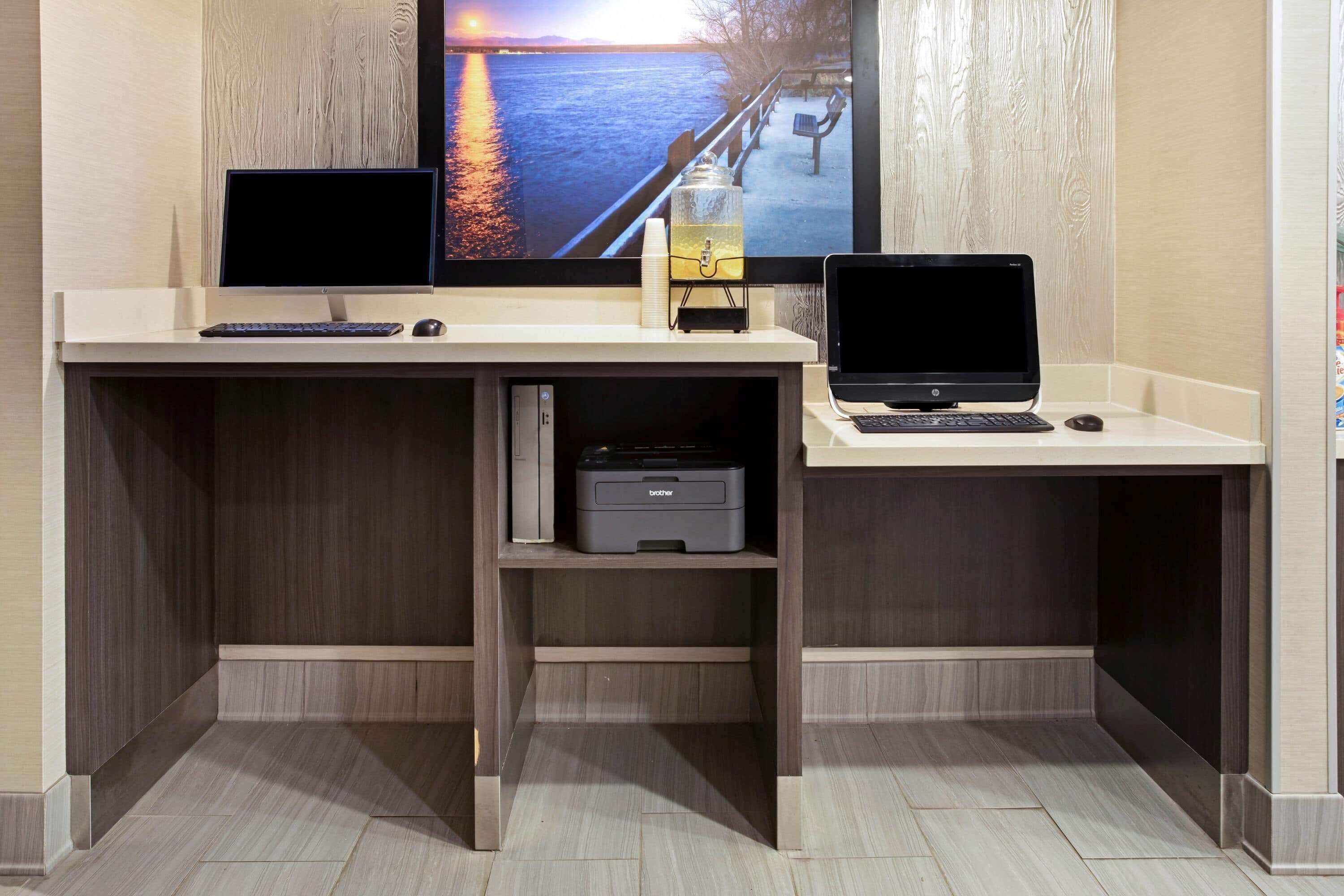 A modern office space with two desks, each with a laptop and a monitor. There is a printer on the left desk, and a water dispenser on the right.