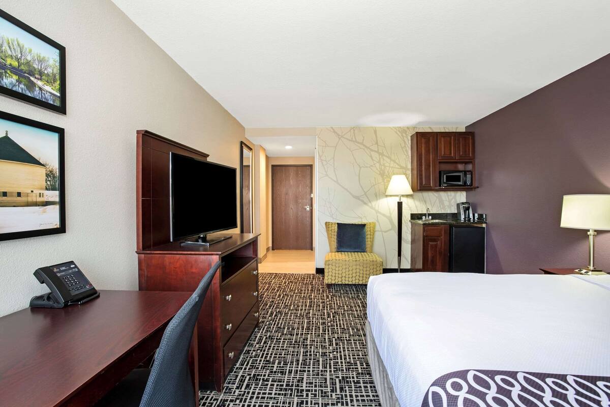 A hotel room with a bed, desk, chair, TV, phone, microwave, and a small kitchenette.