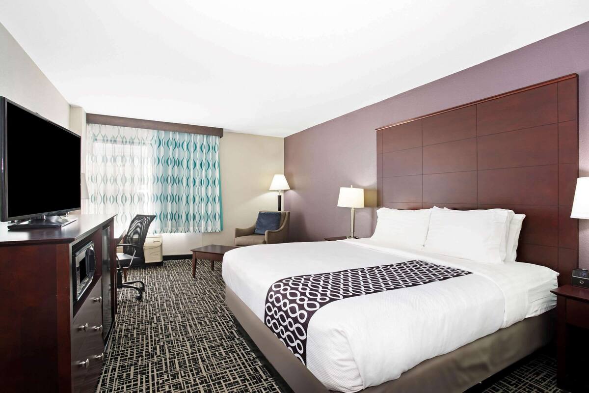 A hotel room with a large bed, a desk with a flat-screen TV, a chair, two bedside lamps, and a window with patterned curtains.