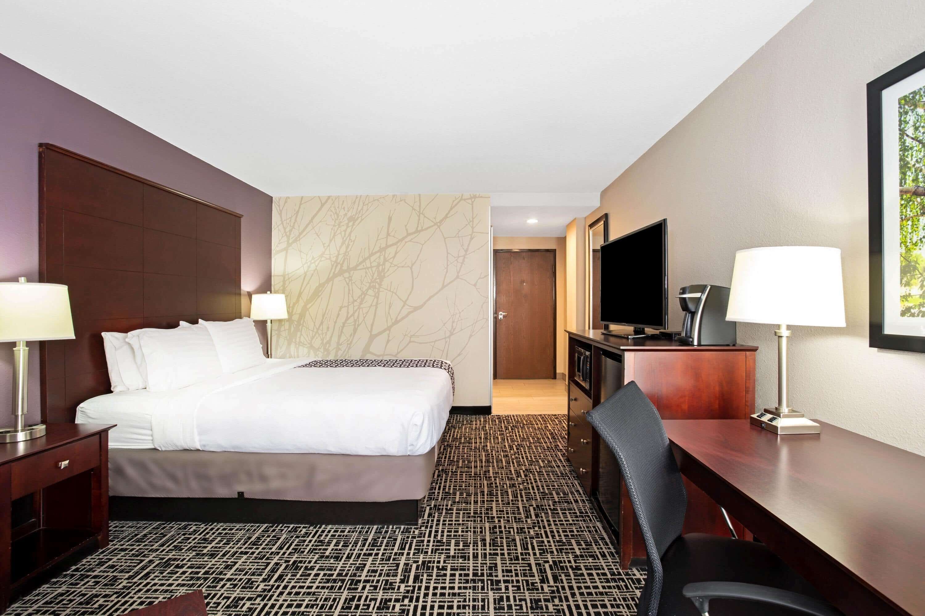 A hotel room with a large bed, two bedside tables with lamps, a desk with a chair, a television, and a door leading to another room.