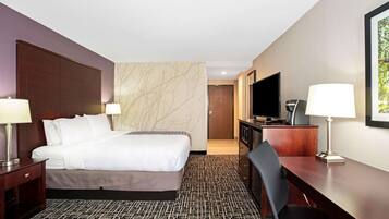 A hotel room with a large bed, two bedside tables with lamps, a desk with a chair, a television, and a door leading to another room.