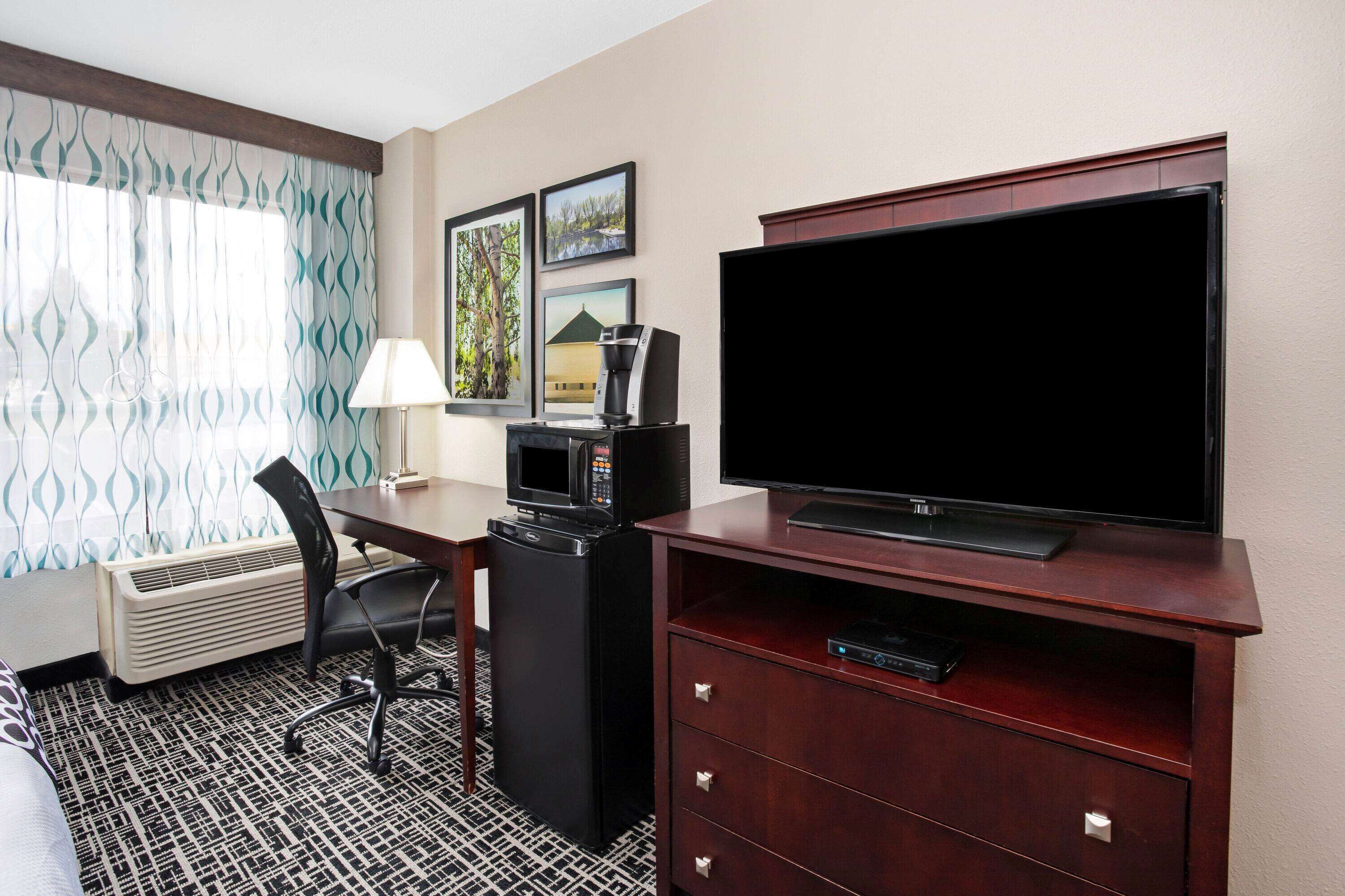 Quarto de hotel com escrivaninha, televisão, micro-ondas, frigorífico, cadeira e tapete estampado.