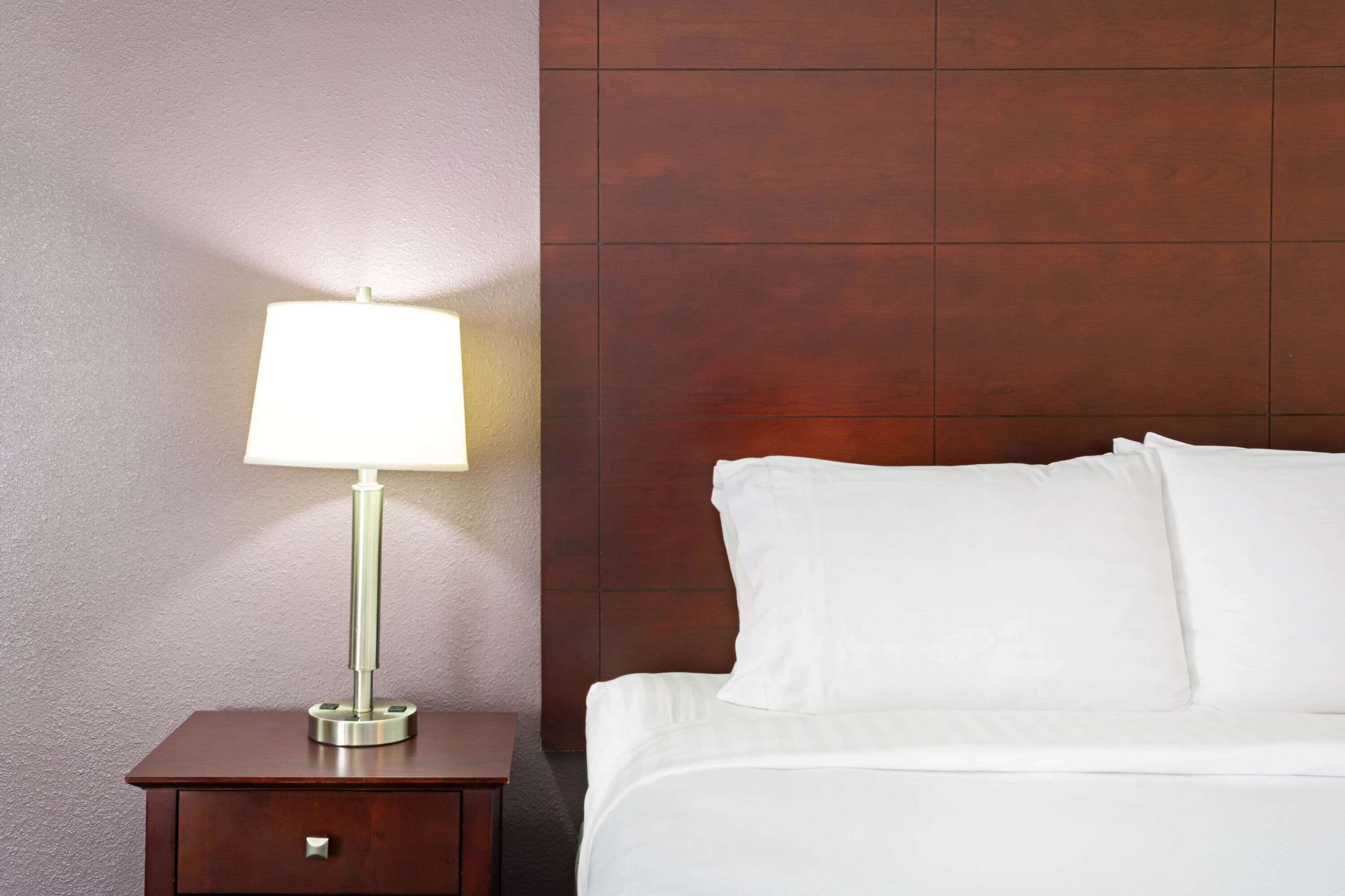 A neatly made bed with white linens and pillows, a bedside table with a lamp, and a wooden headboard.