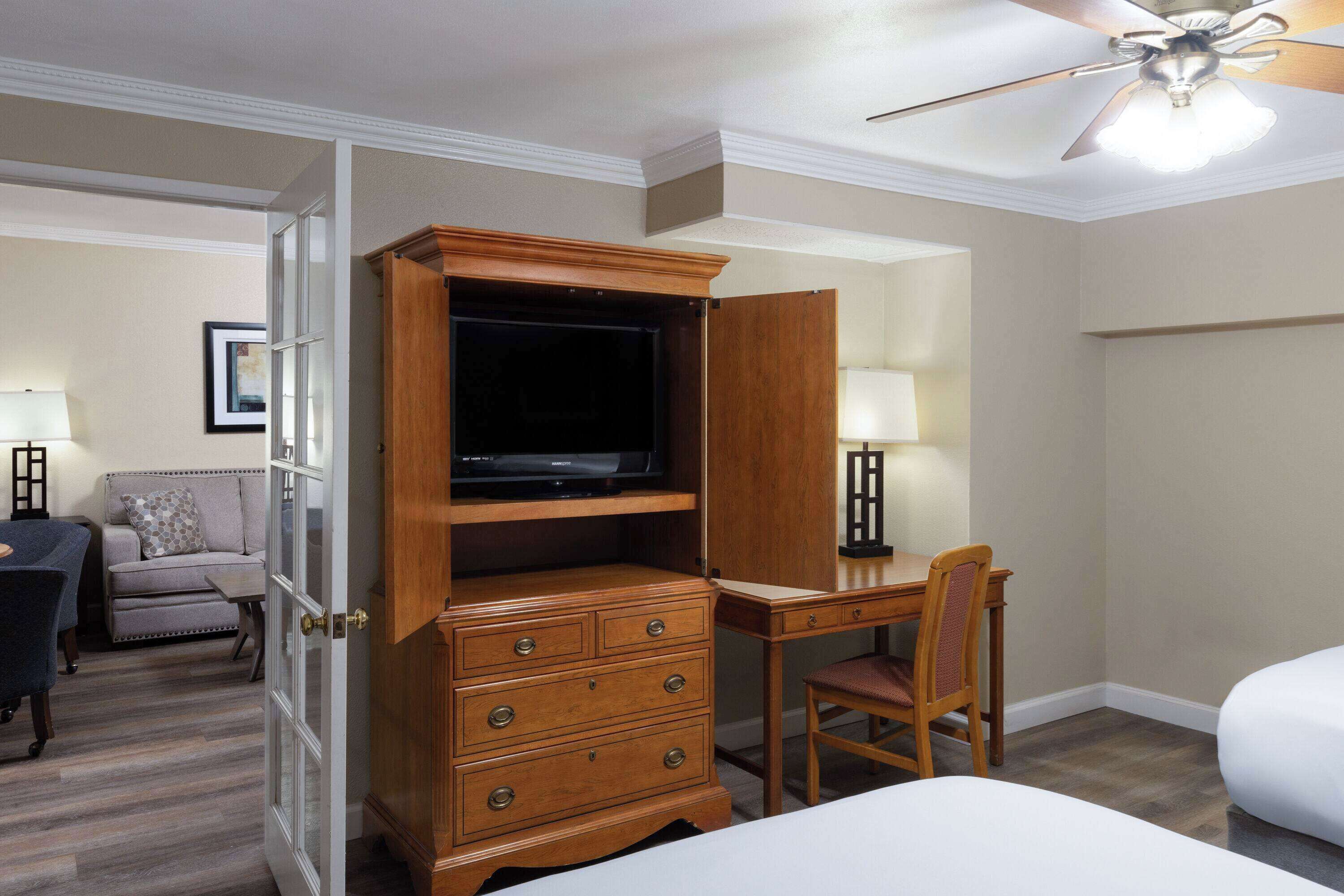 A hotel room with a wooden desk, a television, a chair, a bed, and a ceiling fan.