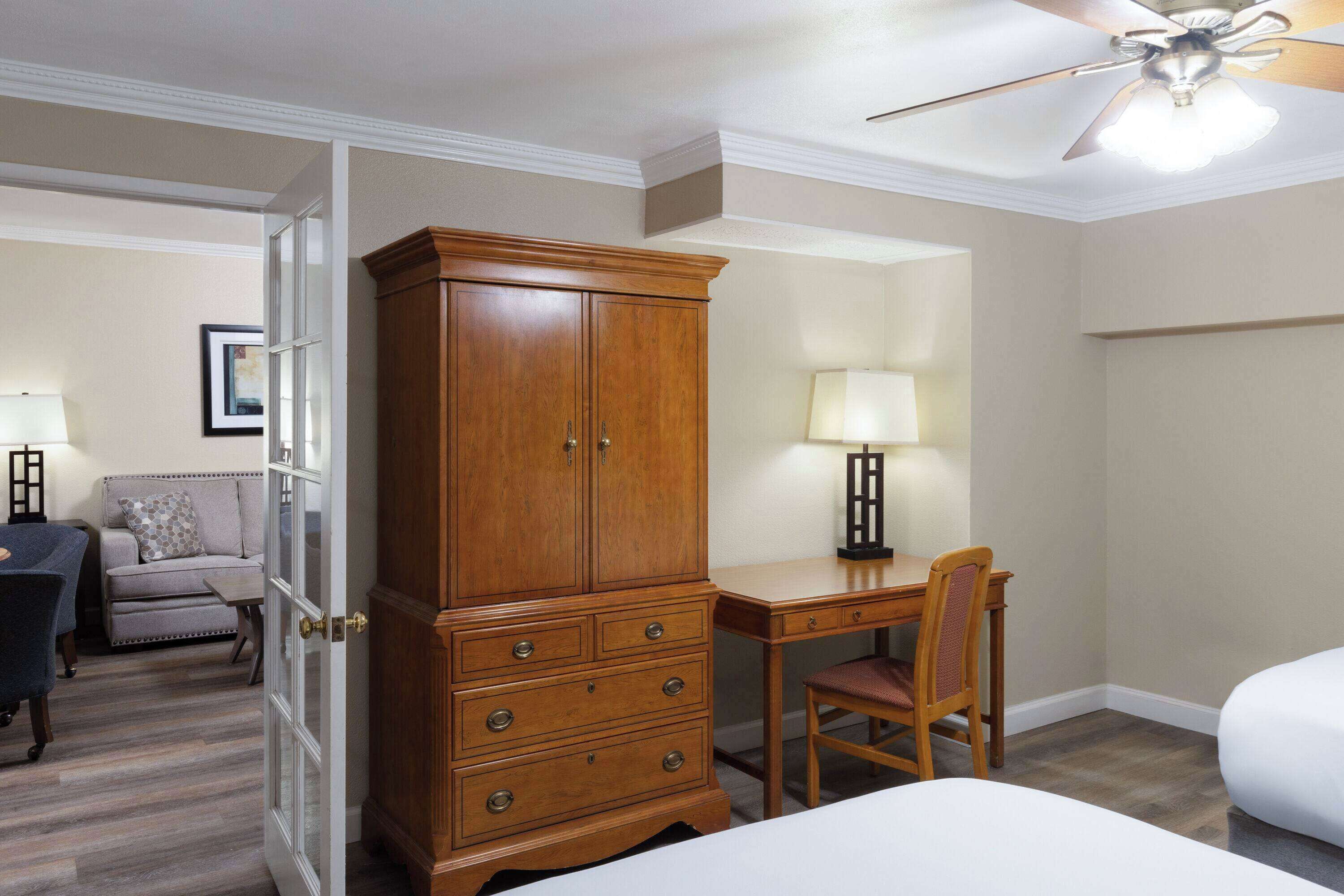 A hotel room with a wooden wardrobe, a desk with a chair, a bed, and a ceiling fan.