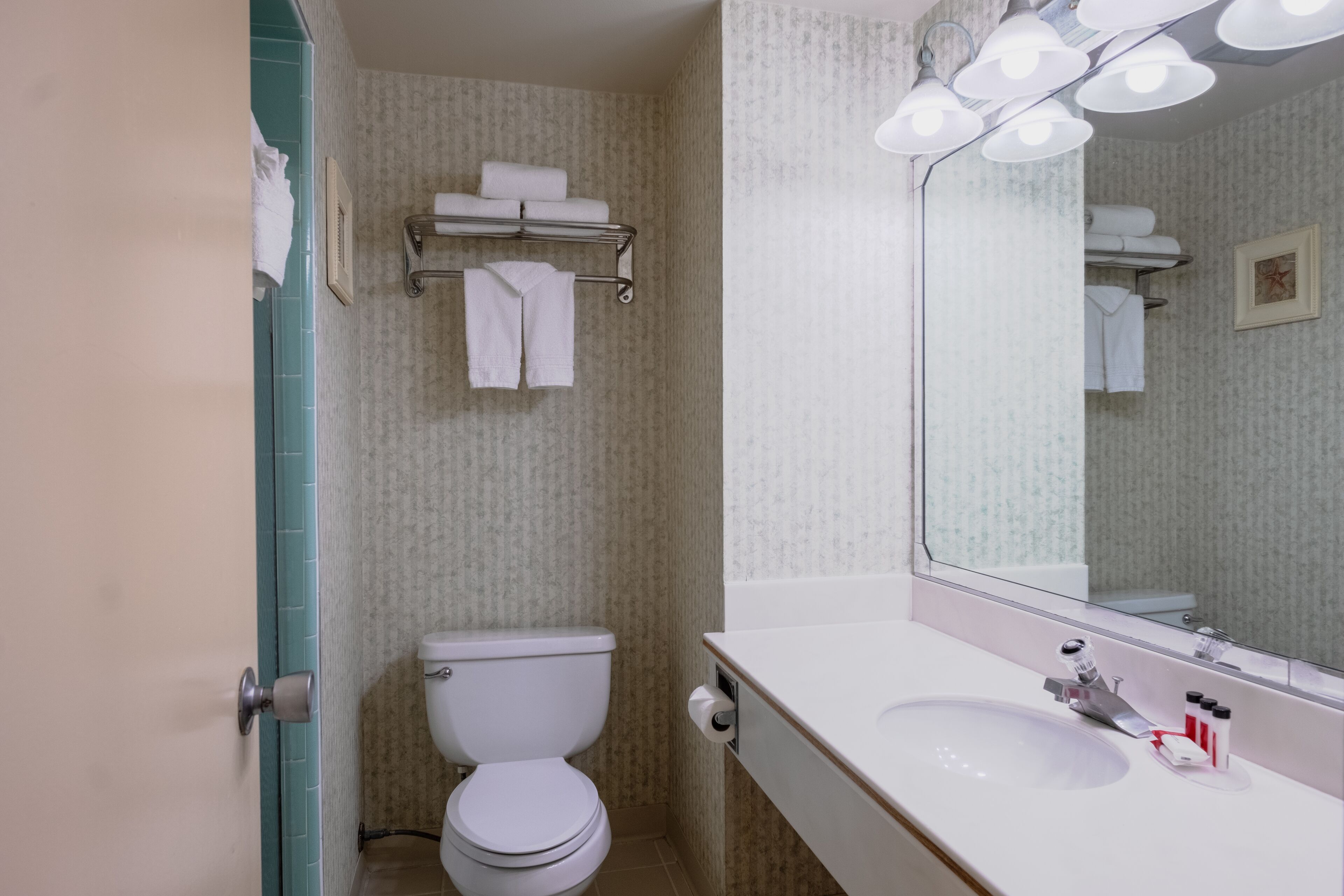 Bany amb vàter, un lavabo i un mirall. Hi ha tovalloles penjades en un escurapulides sobre el vàter.
