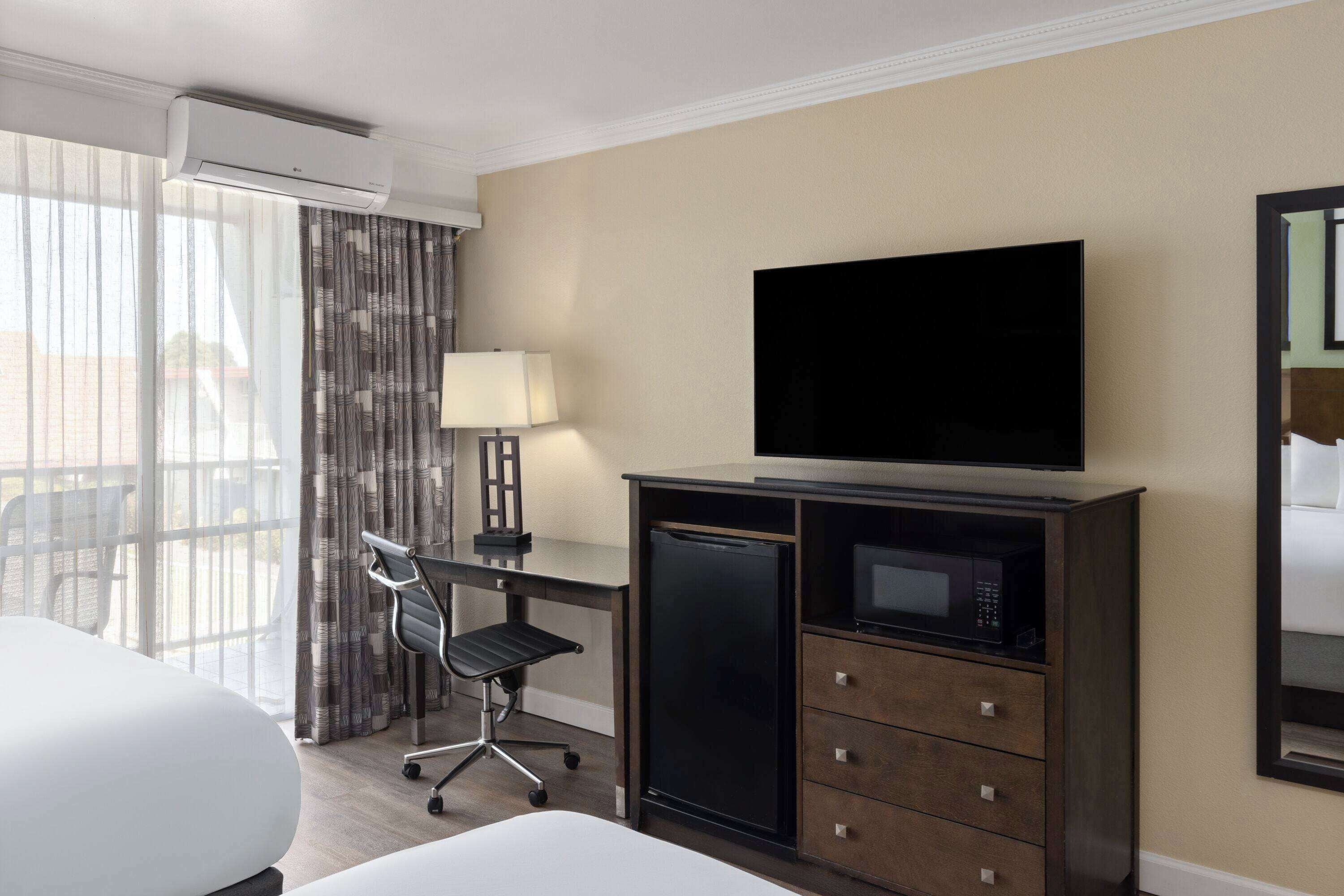 Una habitación de hotel con cama, escritorio con silla, televisor, microondas y un ventanal con cortinas.