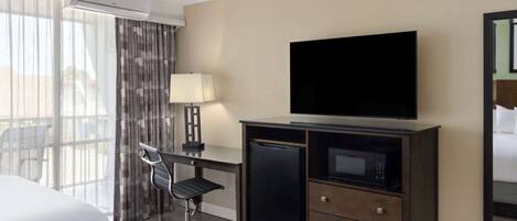 A hotel room with a bed, a desk with a chair, a television, a microwave, and a large window with curtains.