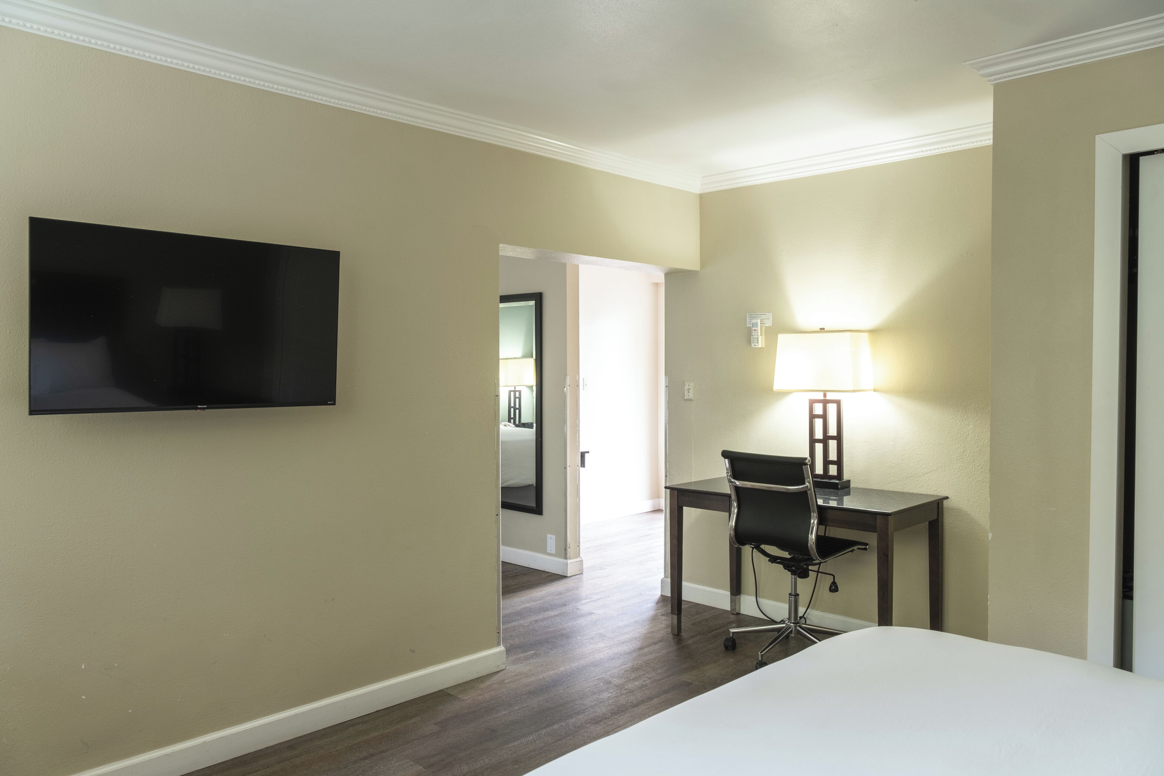 A hotel room with a bed, a desk with a chair, a TV mounted on the wall, and a lamp on the desk.