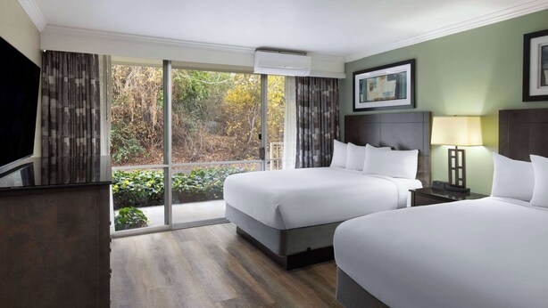 Une chambre d’hôtel avec deux lits, une télévision, une lampe et une vue sur les arbres à travers de grandes fenêtres.