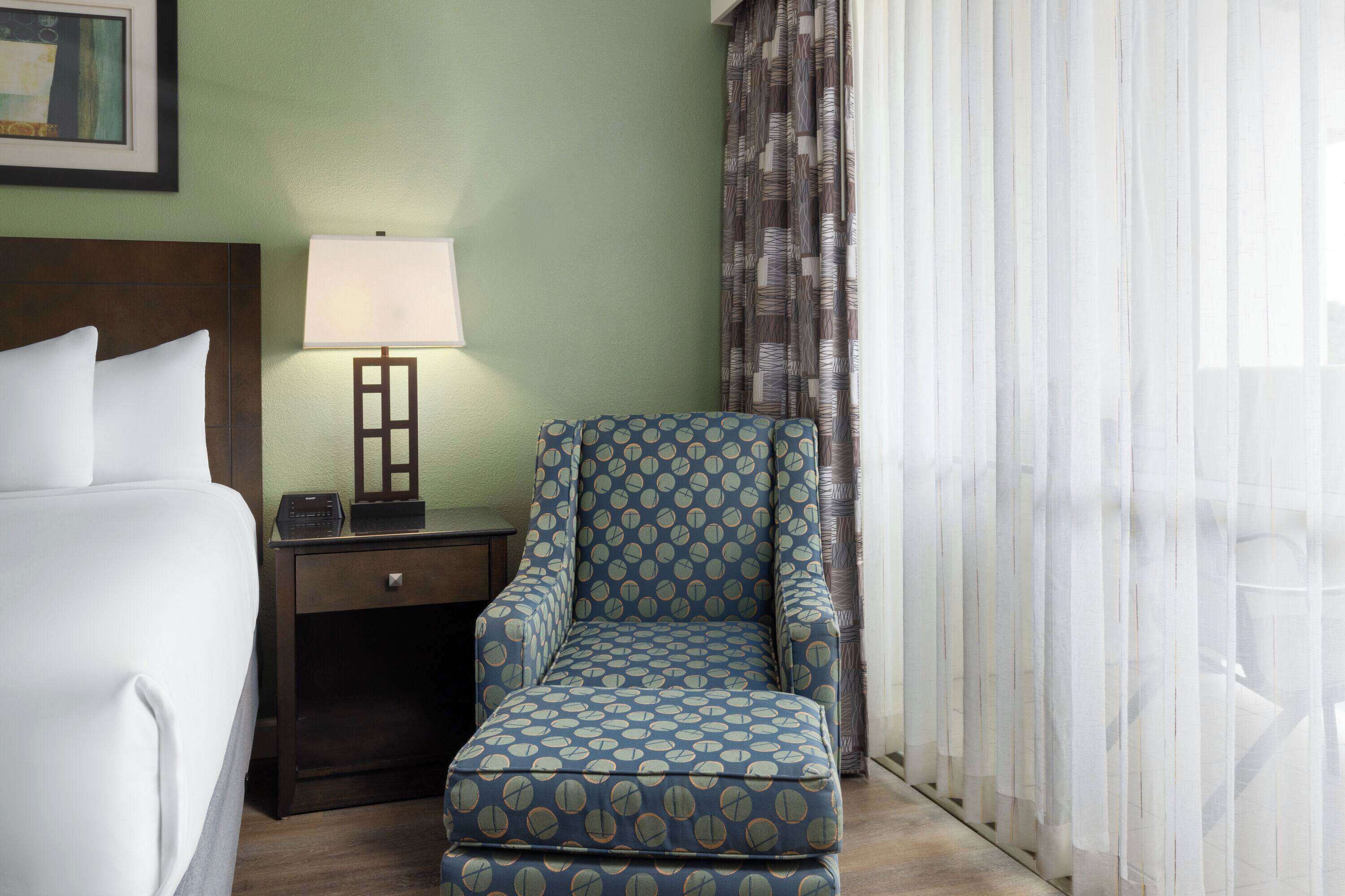 A hotel room with a bed, a nightstand with a lamp, a patterned armchair, and sheer curtains.