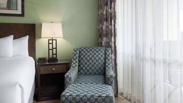 A hotel room with a bed, a nightstand with a lamp, a patterned armchair, and sheer curtains.