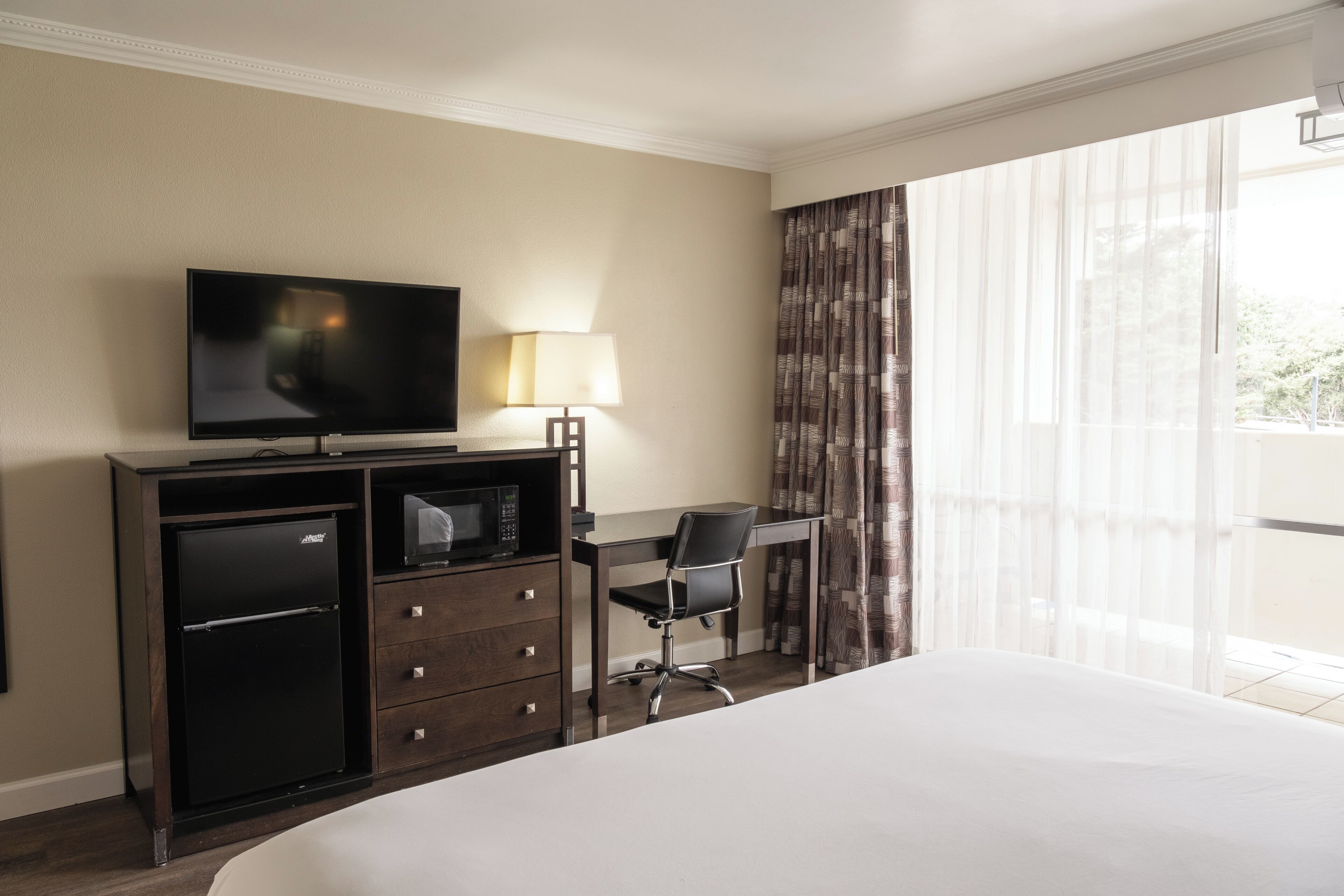 A hotel room with a bed, a desk, a television, a refrigerator, and a lamp.