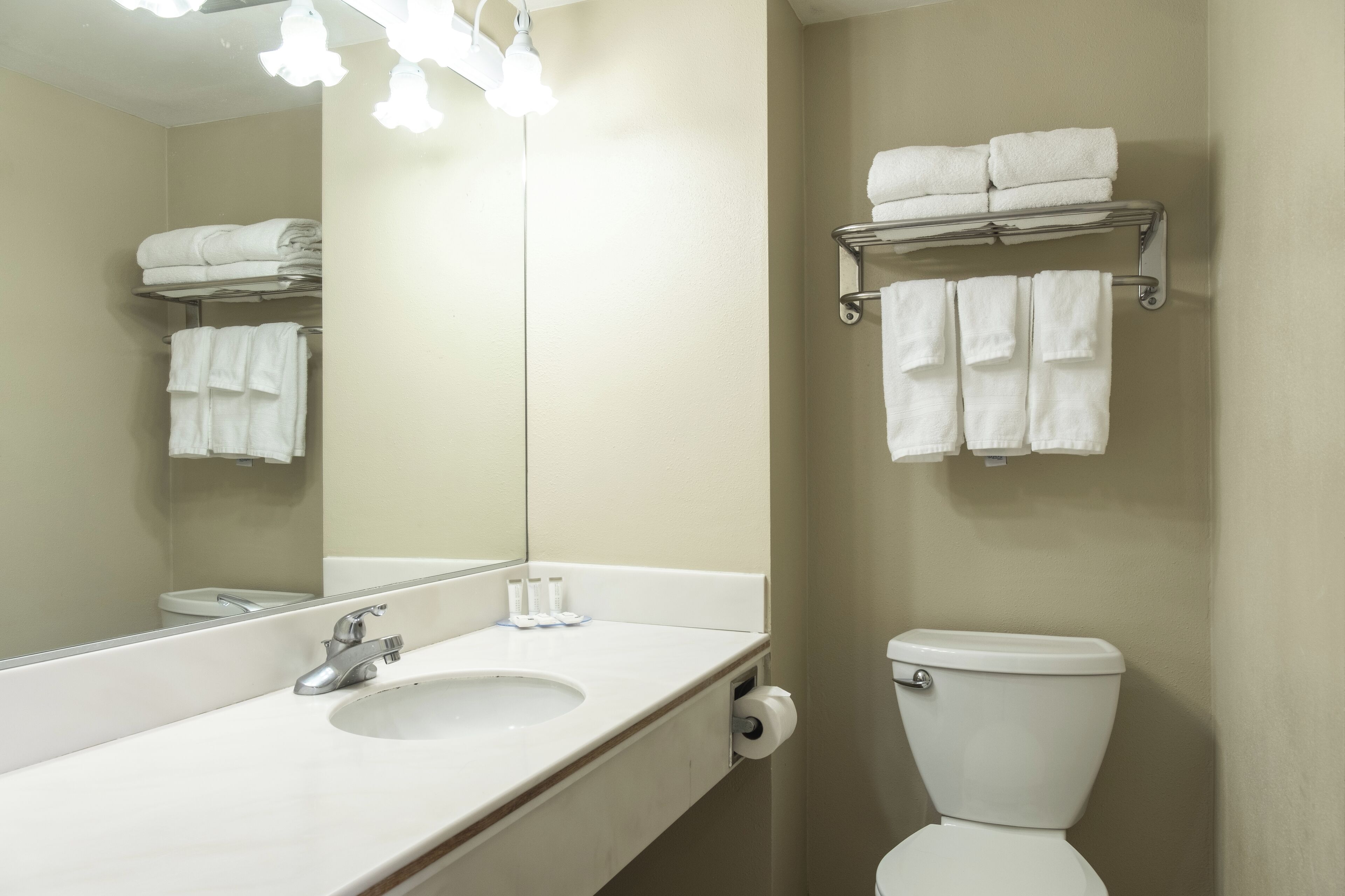 Une salle de bain avec un lavabo, un miroir et des toilettes. Il y a des serviettes sur un porte-serviettes au-dessus des toilettes et une étagère avec des serviettes au-dessus du lavabo.