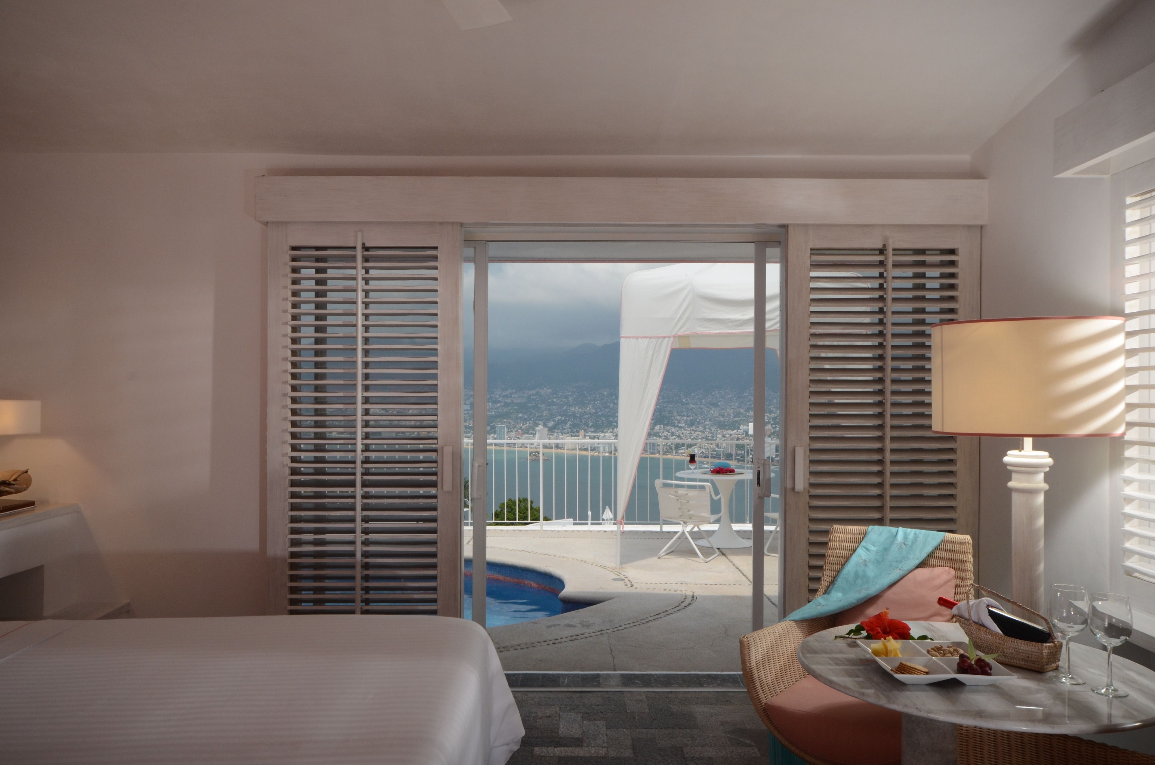 Ein Hotelzimmer mit Bett, Tisch mit Korb und Weingläsern, Balkon mit Blick auf Stadt und Berge sowie ein Schwimmbad.