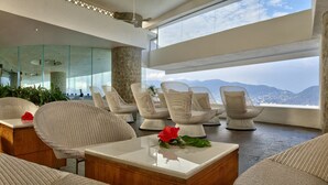 Un salon moderne avec des fauteuils en osier, une table basse agrémentée d’un bouquet de fleurs et une vue sur les montagnes, visible à travers de grandes fenêtres.