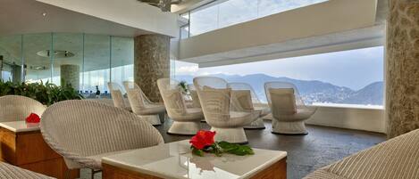 A modern lounge with wicker chairs, a coffee table with a flower, and a view of mountains through large windows.