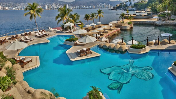 Une grande piscine d’un bleu éclatant, ornée d’un motif floral, entourée de chaises longues et de parasols, et offrant une vue sur la mer et le paysage urbain.