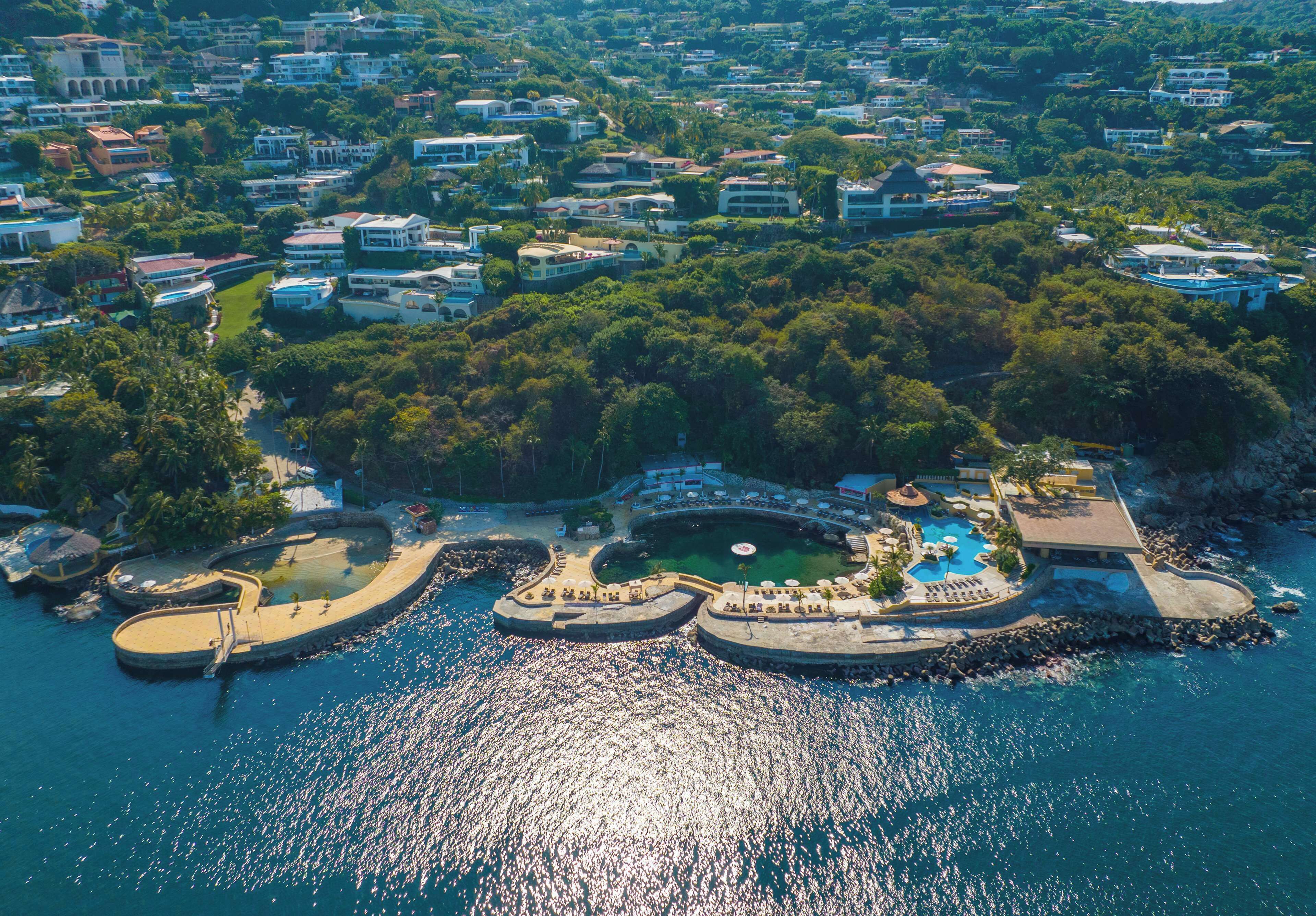 An aerial view of a coastal resort with multiple villas, a central pool area, and surrounding greenery.