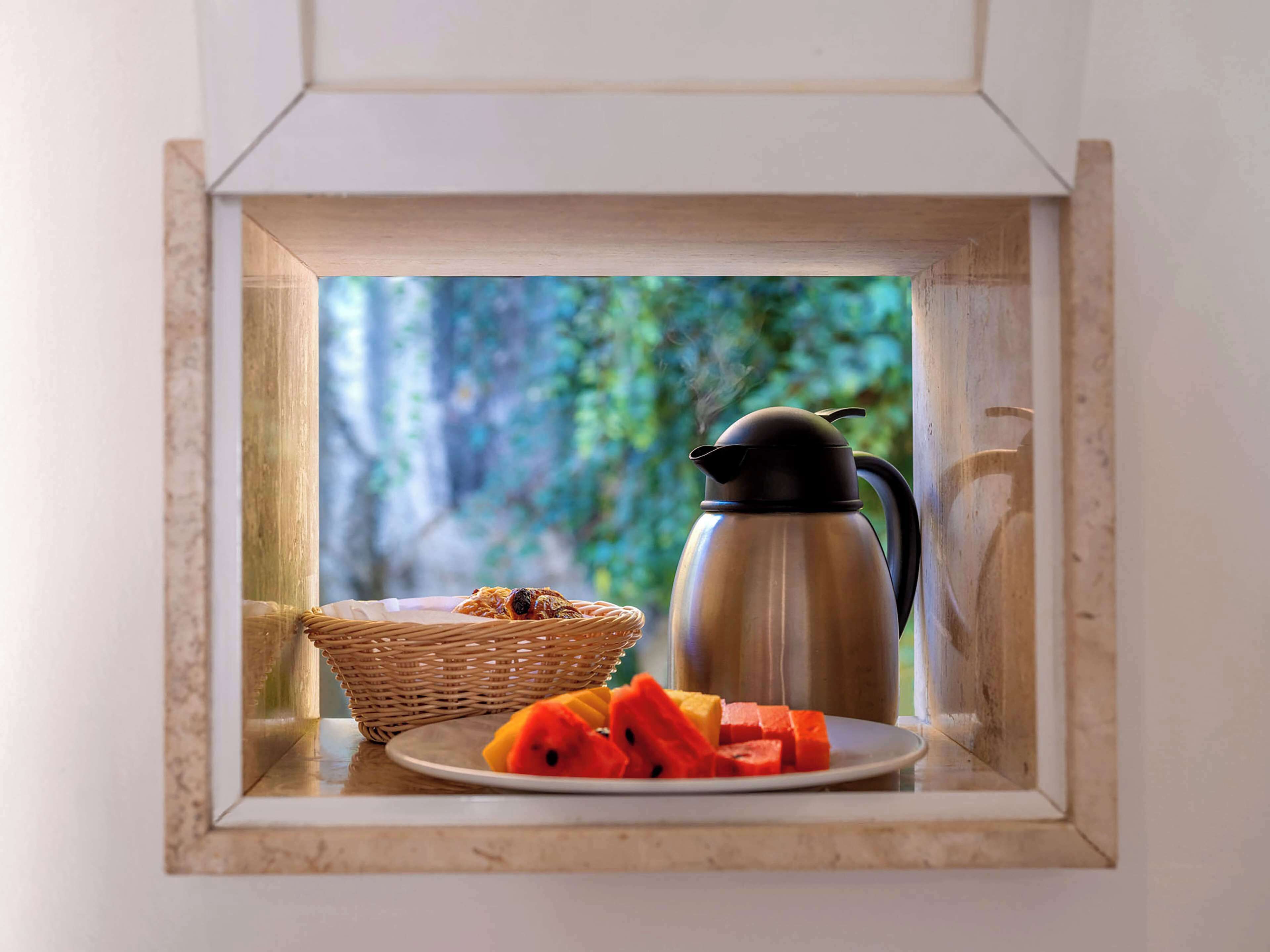 Mesa de café da manhã com uma cafeteira de aço inoxidável, um prato de frutas fatiadas e uma cesta de vime em um parapeito de janela.