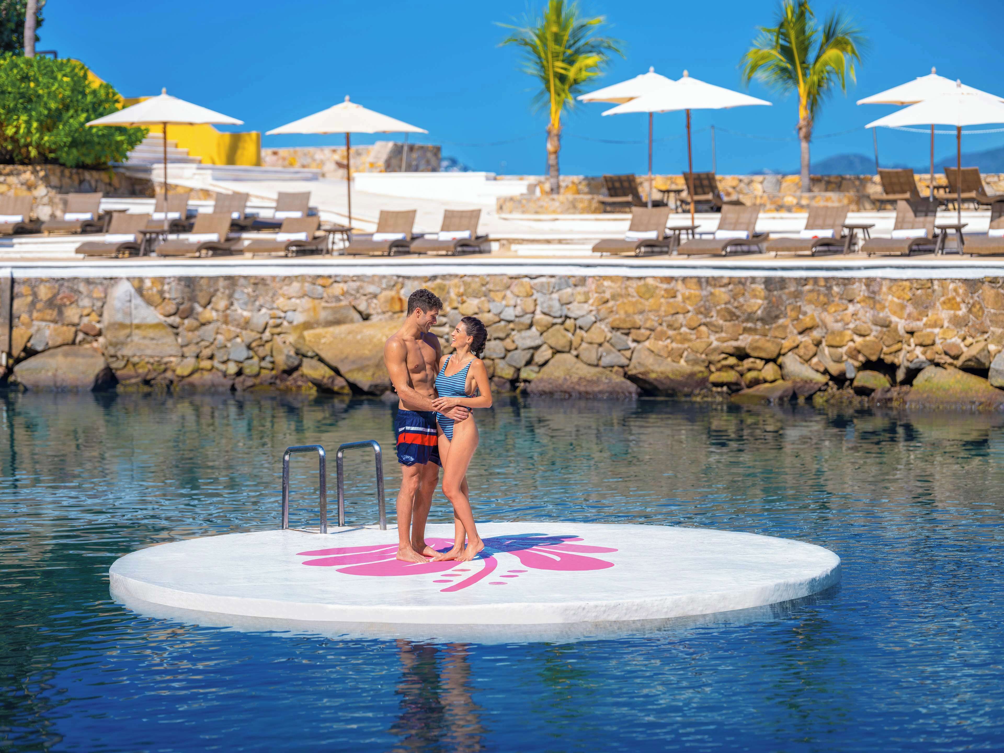 A couple standing on a circular platform in a pool with lounge chairs and umbrellas in the background.