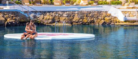 A pool area with a couple sitting on a floating platform, surrounded by lounge chairs and umbrellas.