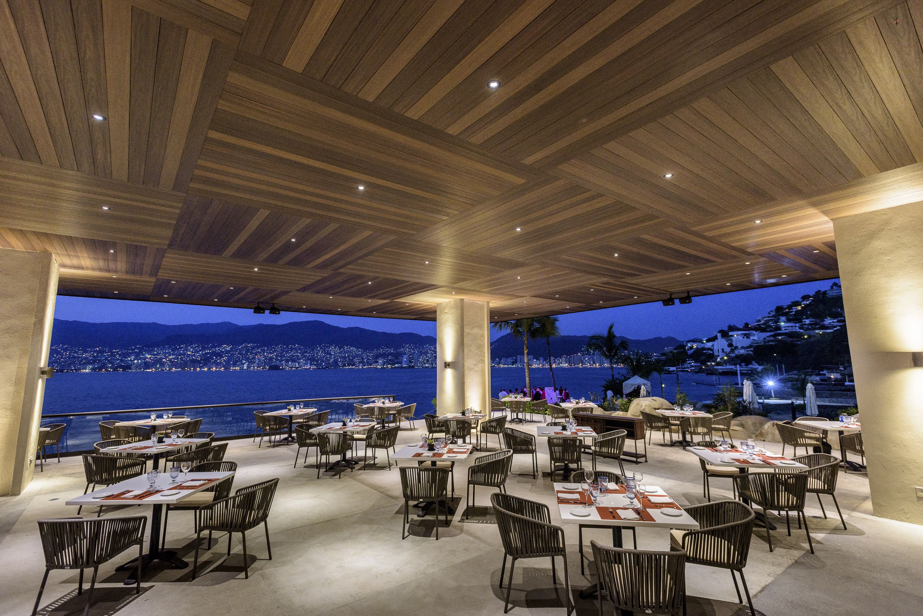 An outdoor dining area with tables and chairs, a wooden ceiling, and a view of the city and mountains at dusk.