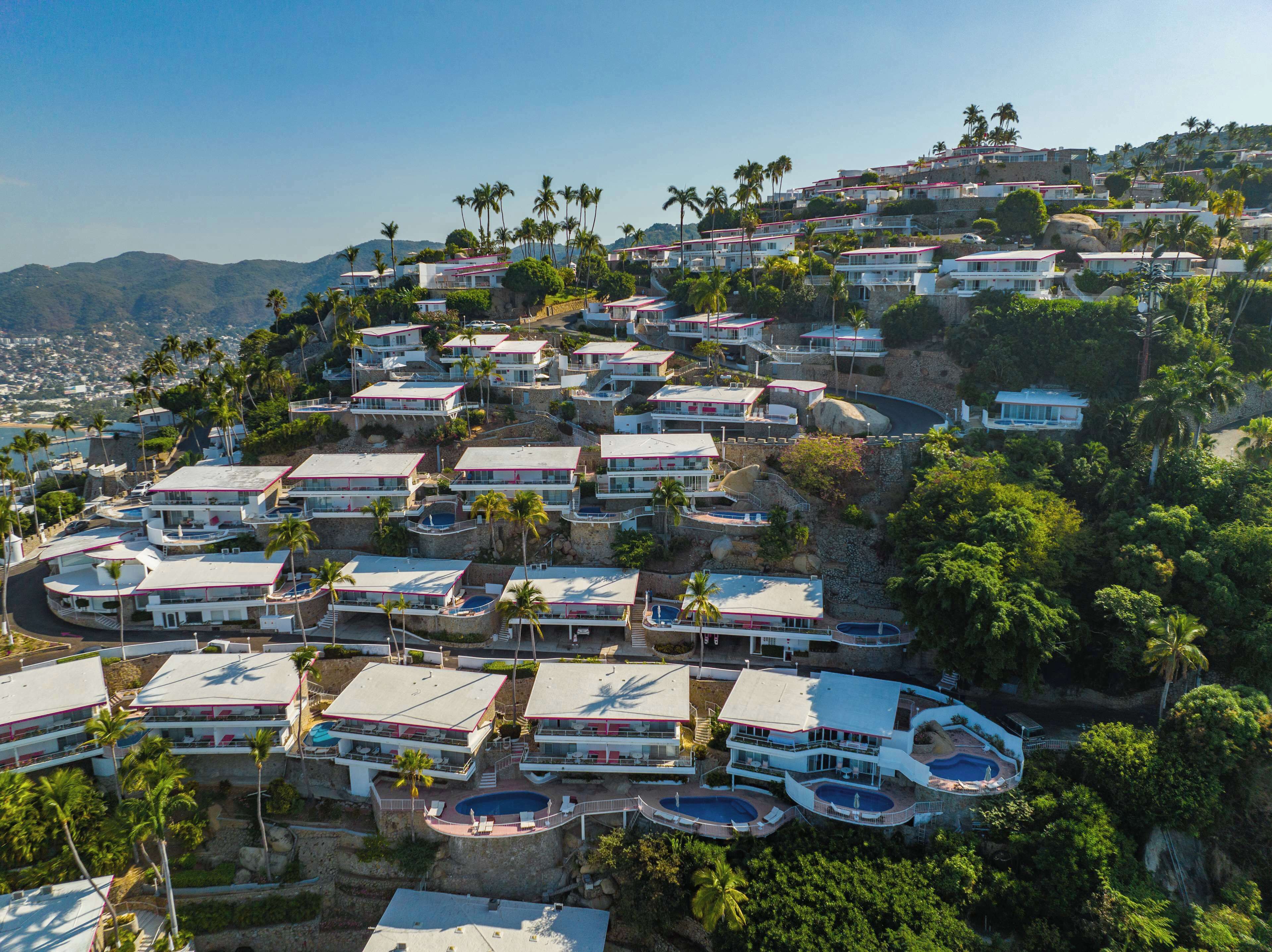 Zona residencial costera con varias casas de dos pisos, balcones, piscinas y abundante vegetación.