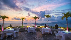 Un espace de restauration en plein air avec des tables et des chaises, des palmiers et une vue sur la mer au coucher du soleil.