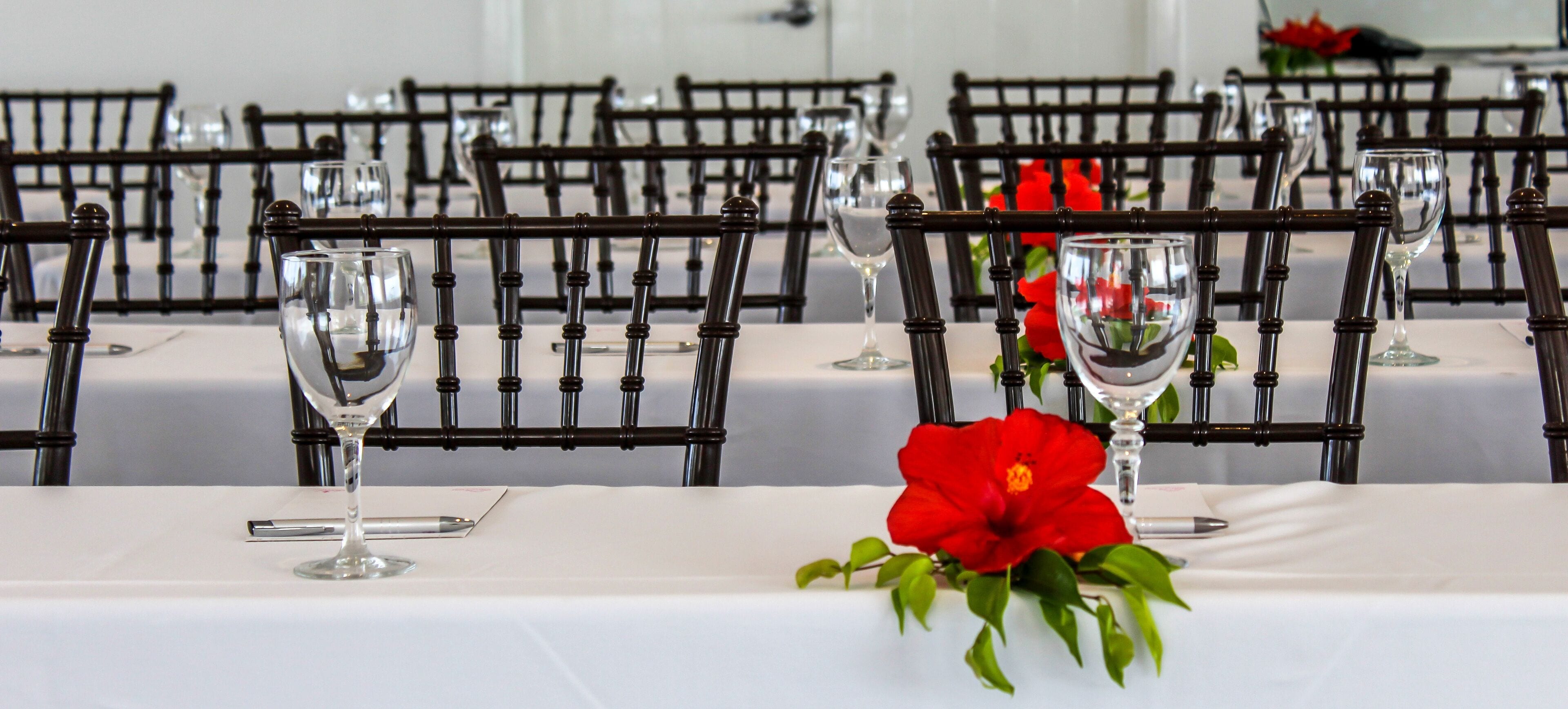A dining table with chairs and wine glasses, a red flower centerpiece, and a white tablecloth.