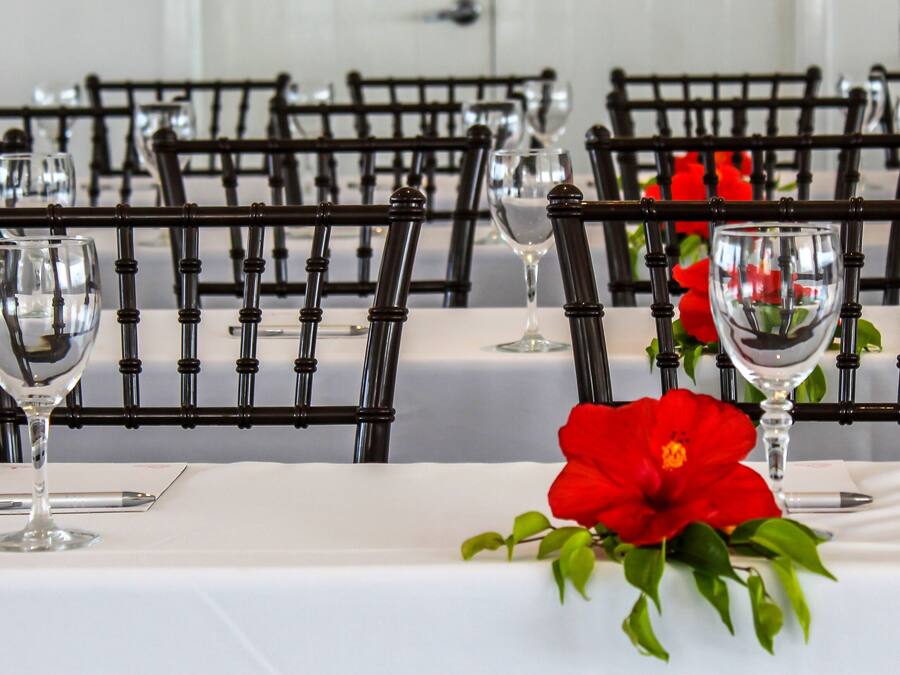 Una mesa de comedor con sillas, copas de vino, un centro de mesa con una flor roja y un mantel blanco.