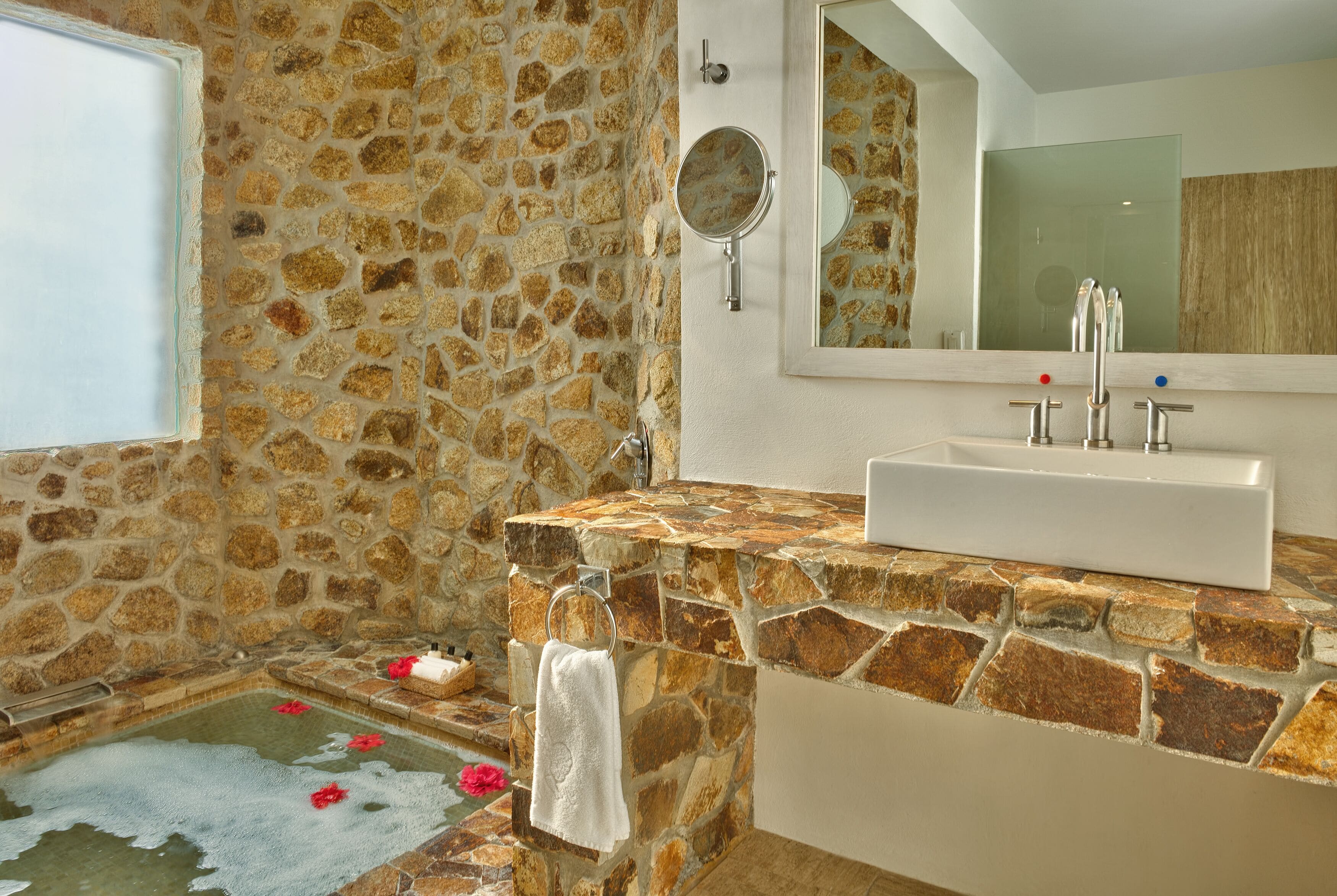 Un baño con pared de piedra, un lavabo, un espejo y una bañera con pétalos de rosa.