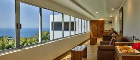A spacious living area with a large window overlooking the ocean, wooden furniture, and a view of a modern building.