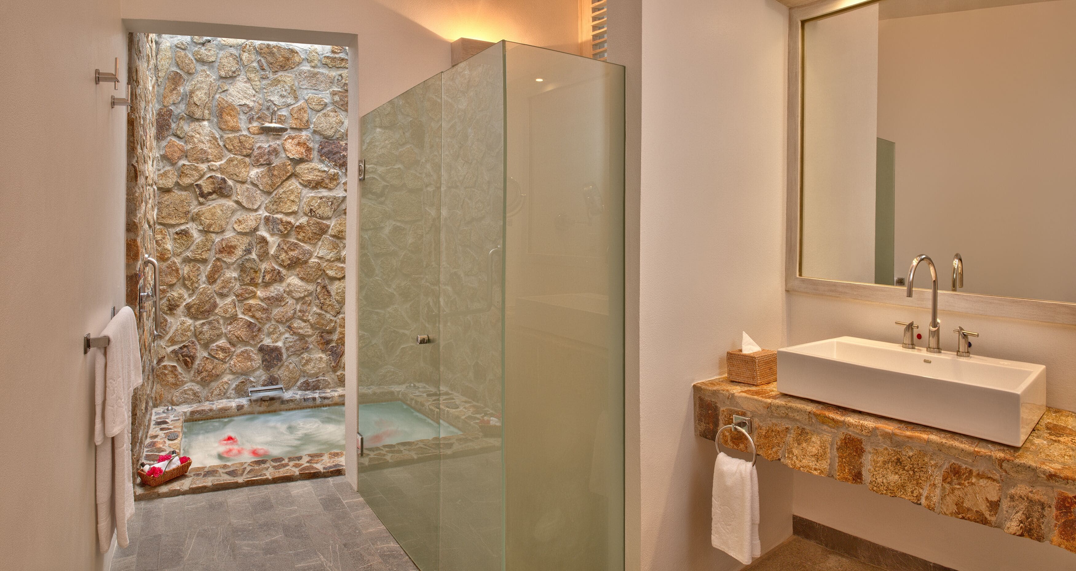 Un baño moderno con ducha de vidrio, pared de piedra, bañera y lavamanos sobre encimera de piedra.