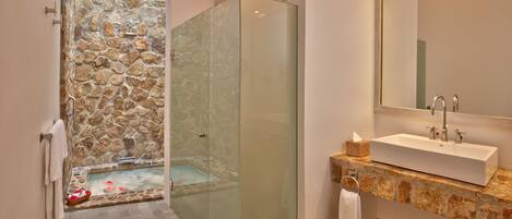 Un baño moderno con ducha de vidrio, pared de piedra, bañera y lavamanos sobre encimera de piedra.