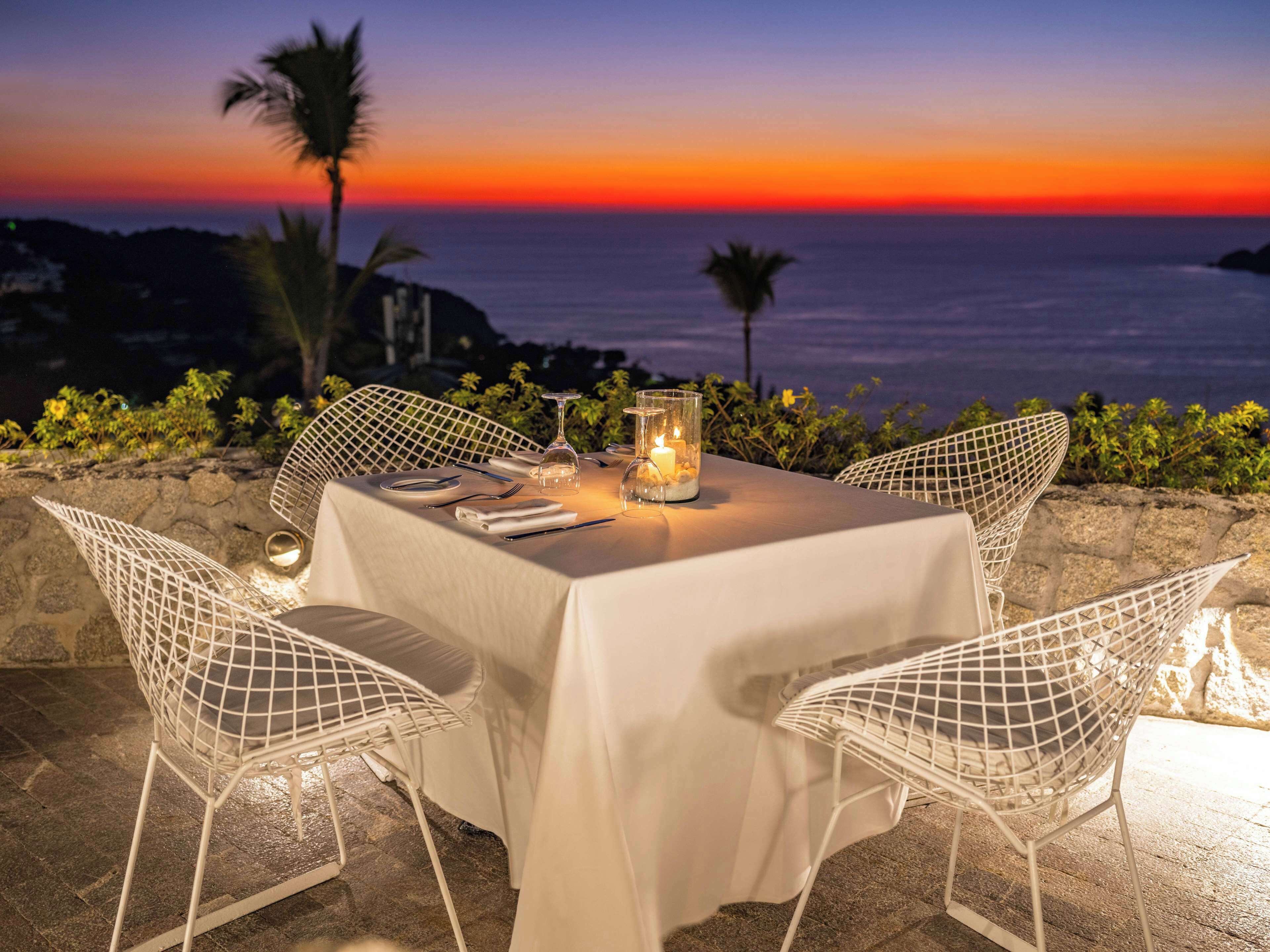 A table set for two with white wireframe chairs, a white tablecloth, and candles, overlooking a sunset over the ocean.