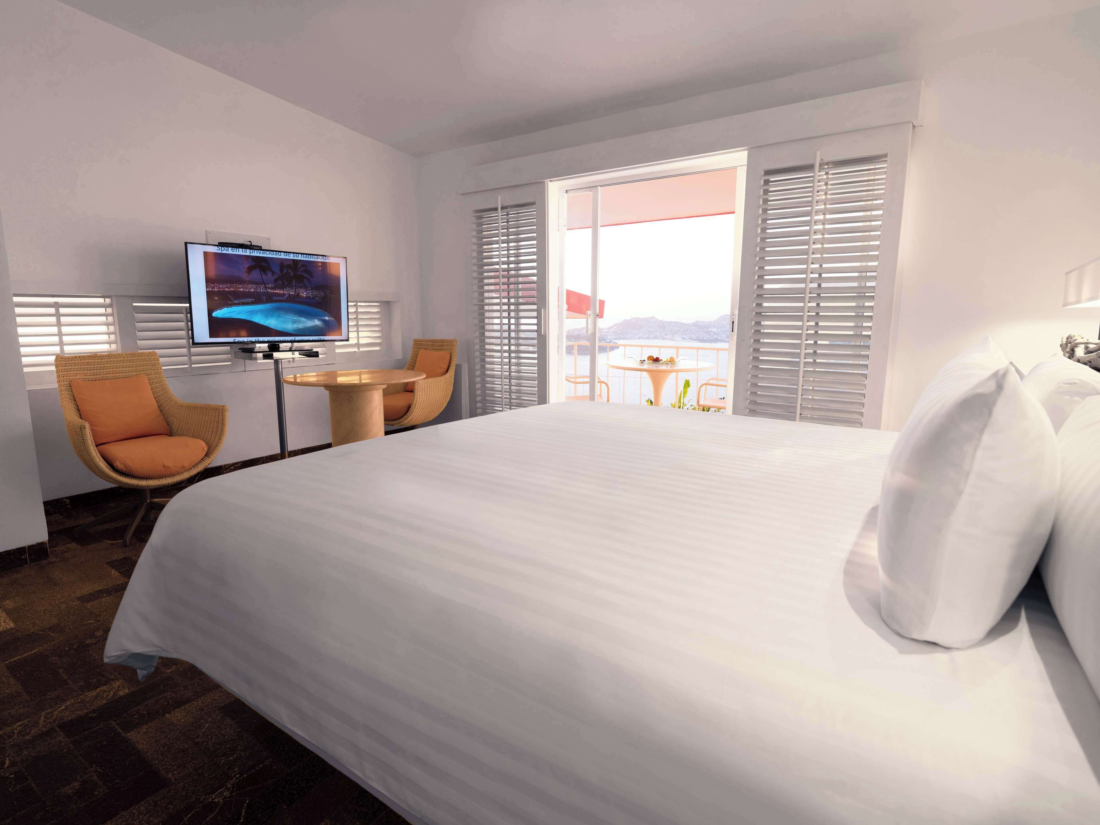 Habitación de hotel con una cama grande, televisión, una silla de mimbre, una mesita redonda y un balcón con vistas al agua.