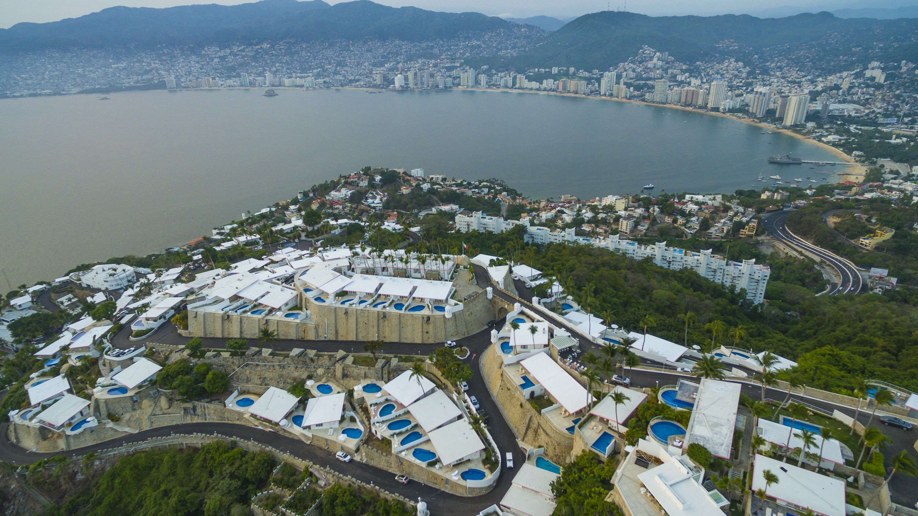An aerial view of a coastal resort with multiple swimming pools, a marina, and a cityscape in the background.