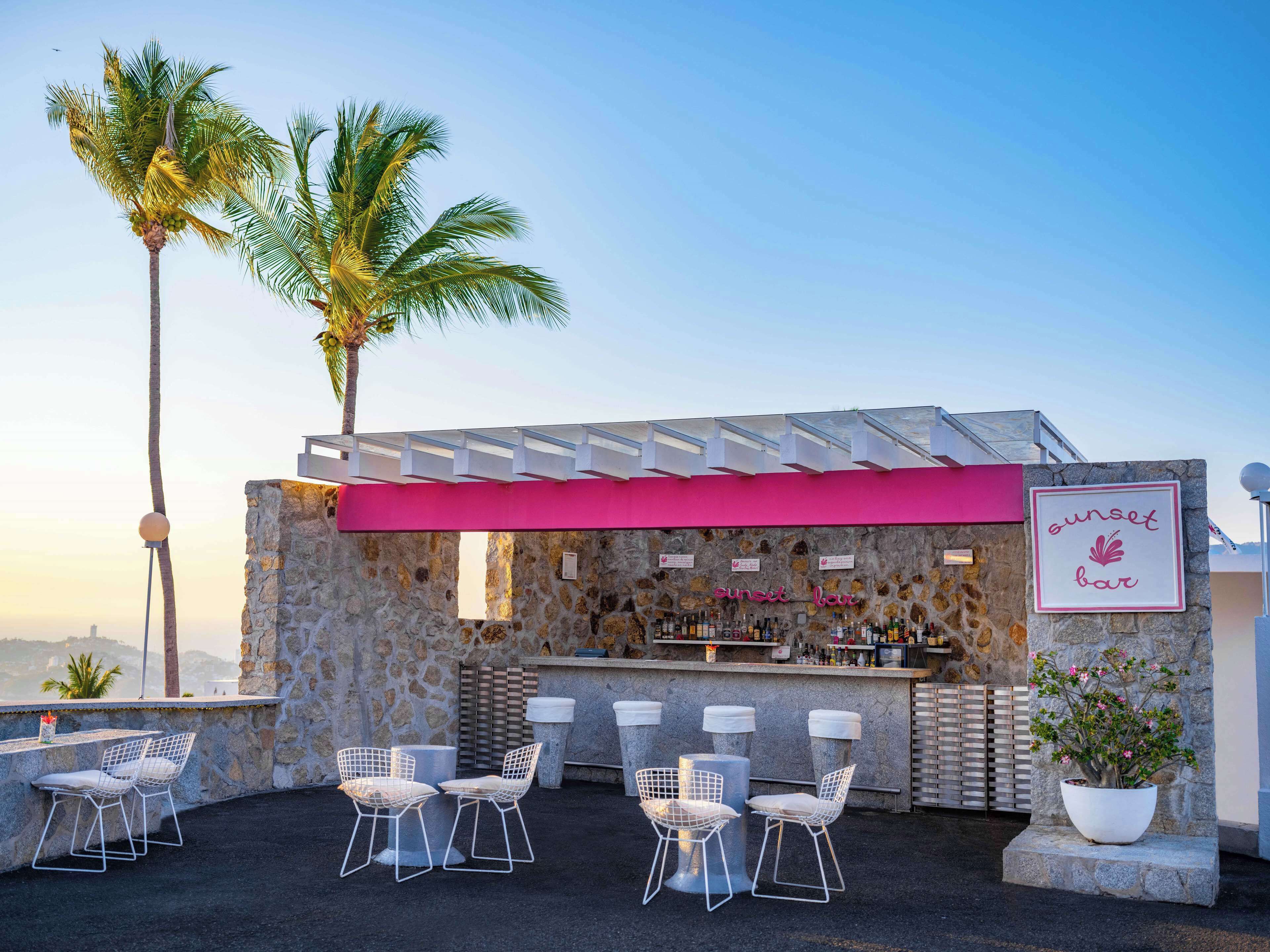 An outdoor bar area with stone walls, a pink awning, and white wire chairs.