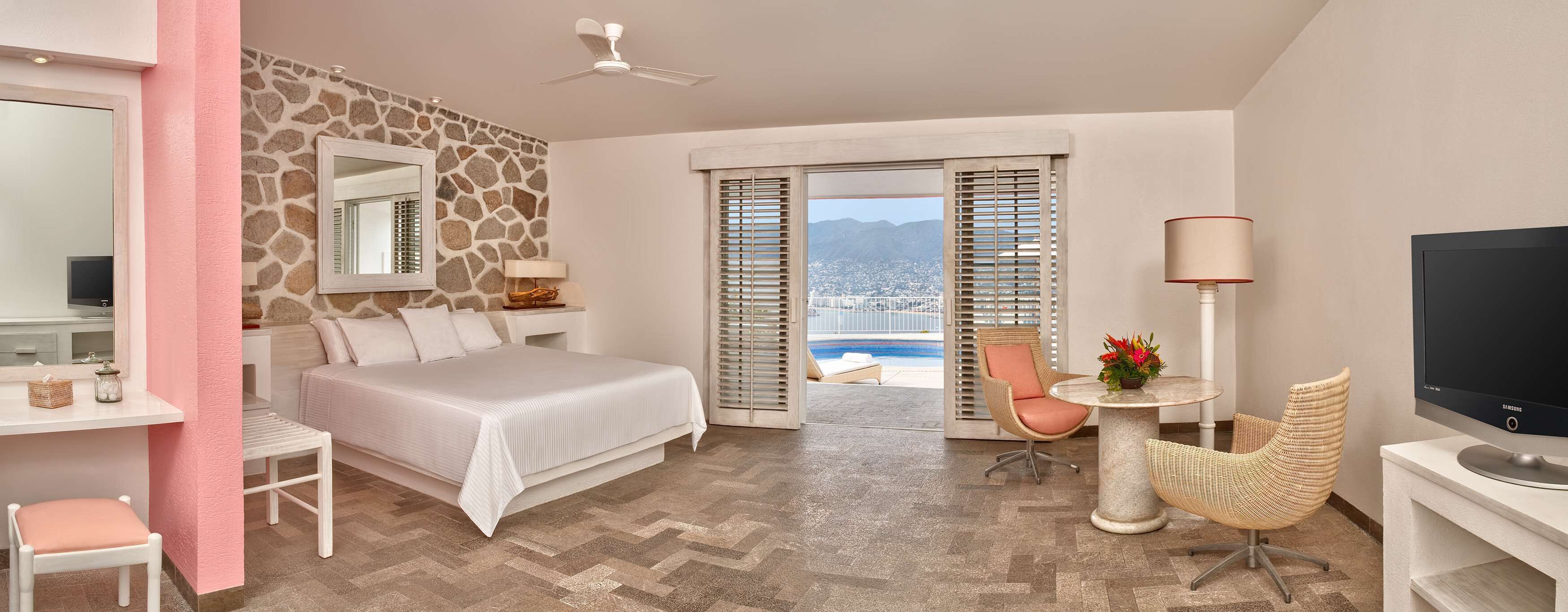 A hotel room with a bed, a desk, a TV, a chair, and a view of a pool and mountains.
