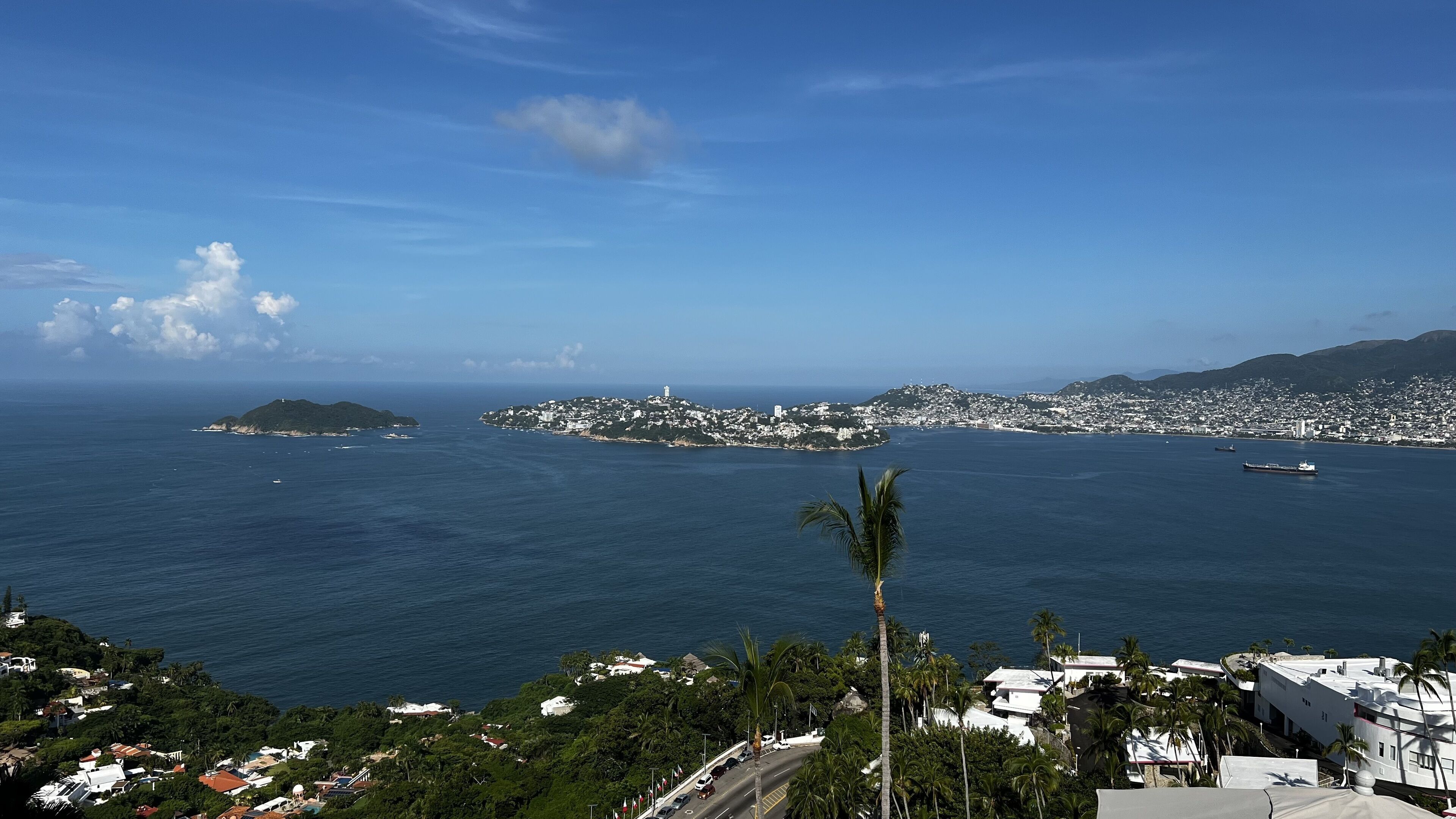 A coastal cityscape with a clear view of the sea, a prominent hill with buildings, and a road leading towards the city.
