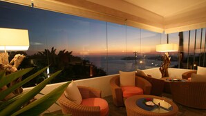 Un balcon agrémenté de meubles en osier, une table en verre ornée d’un centre de table, et une vue sur le coucher de soleil au-dessus de l’eau.