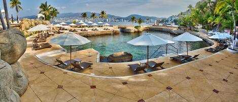 A resort pool area with lounge chairs and umbrellas, a water feature, and a view of the city and mountains in the background.