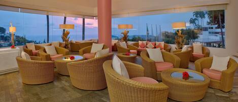 A spacious lounge area with wicker furniture and large windows offering a view of palm trees and a distant cityscape.