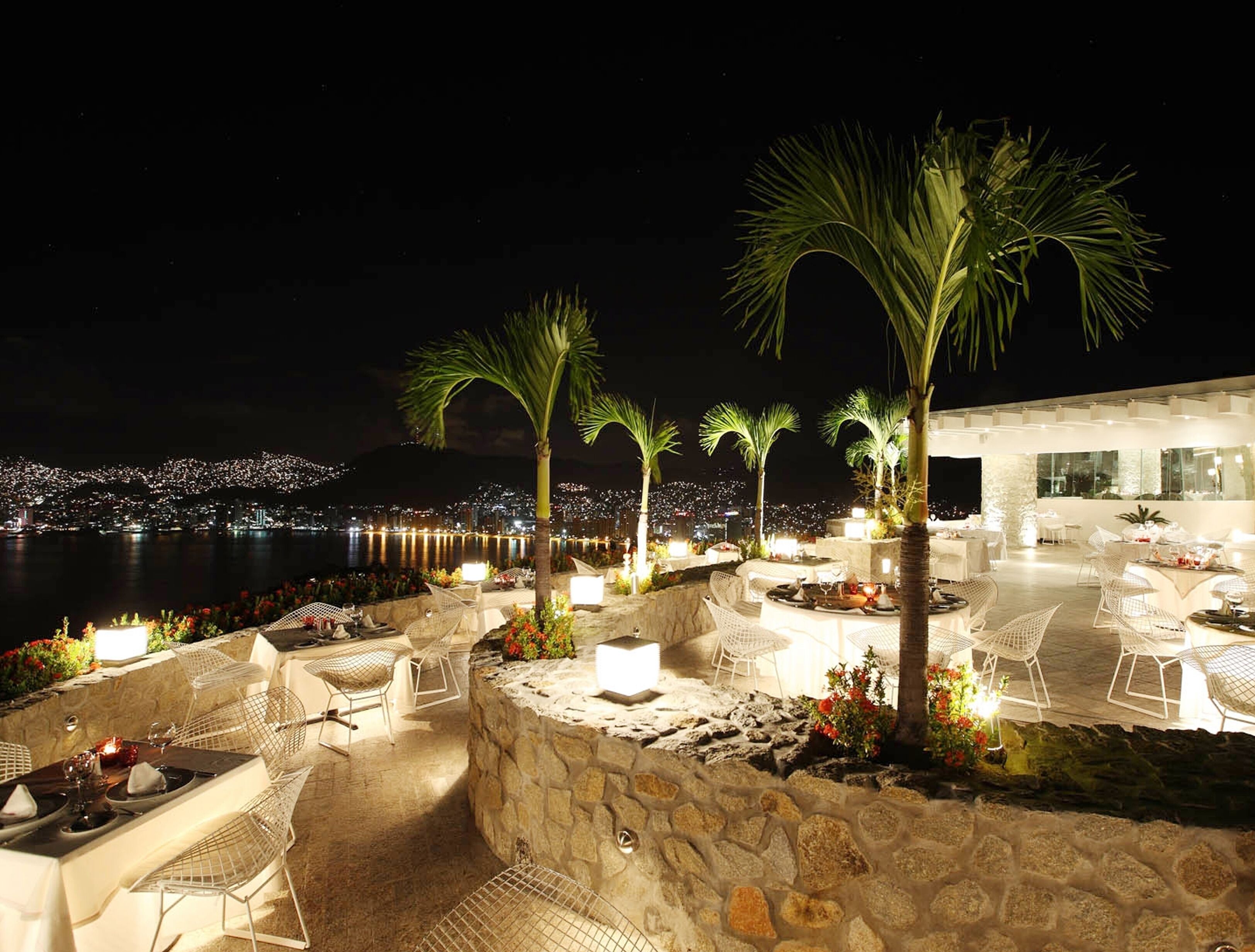 An outdoor dining area with stone walls, white chairs, and palm trees.